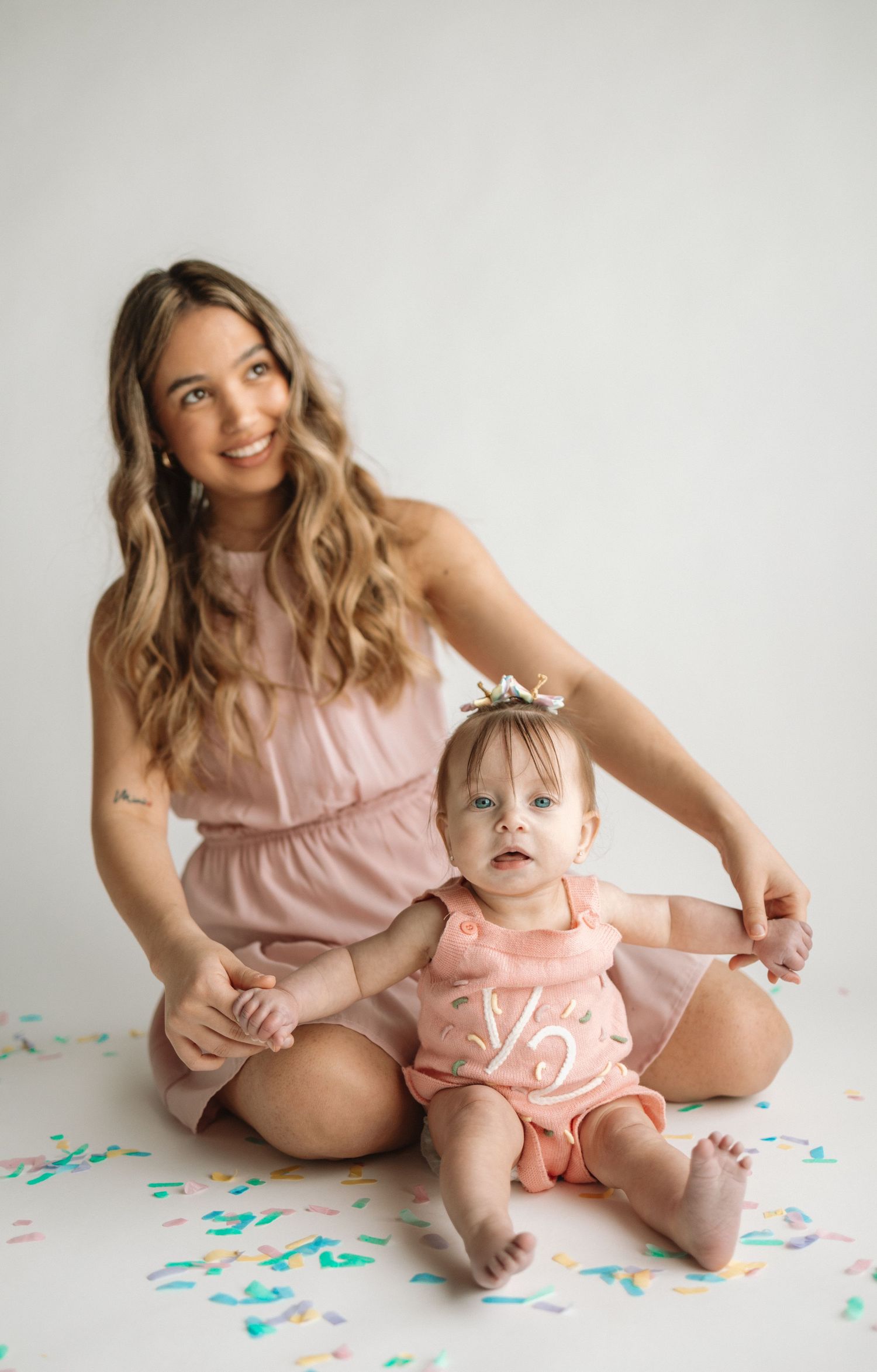 A series of four photos showing a parent and baby in matching pink outfits sitting on a white backdrop with teal confetti.