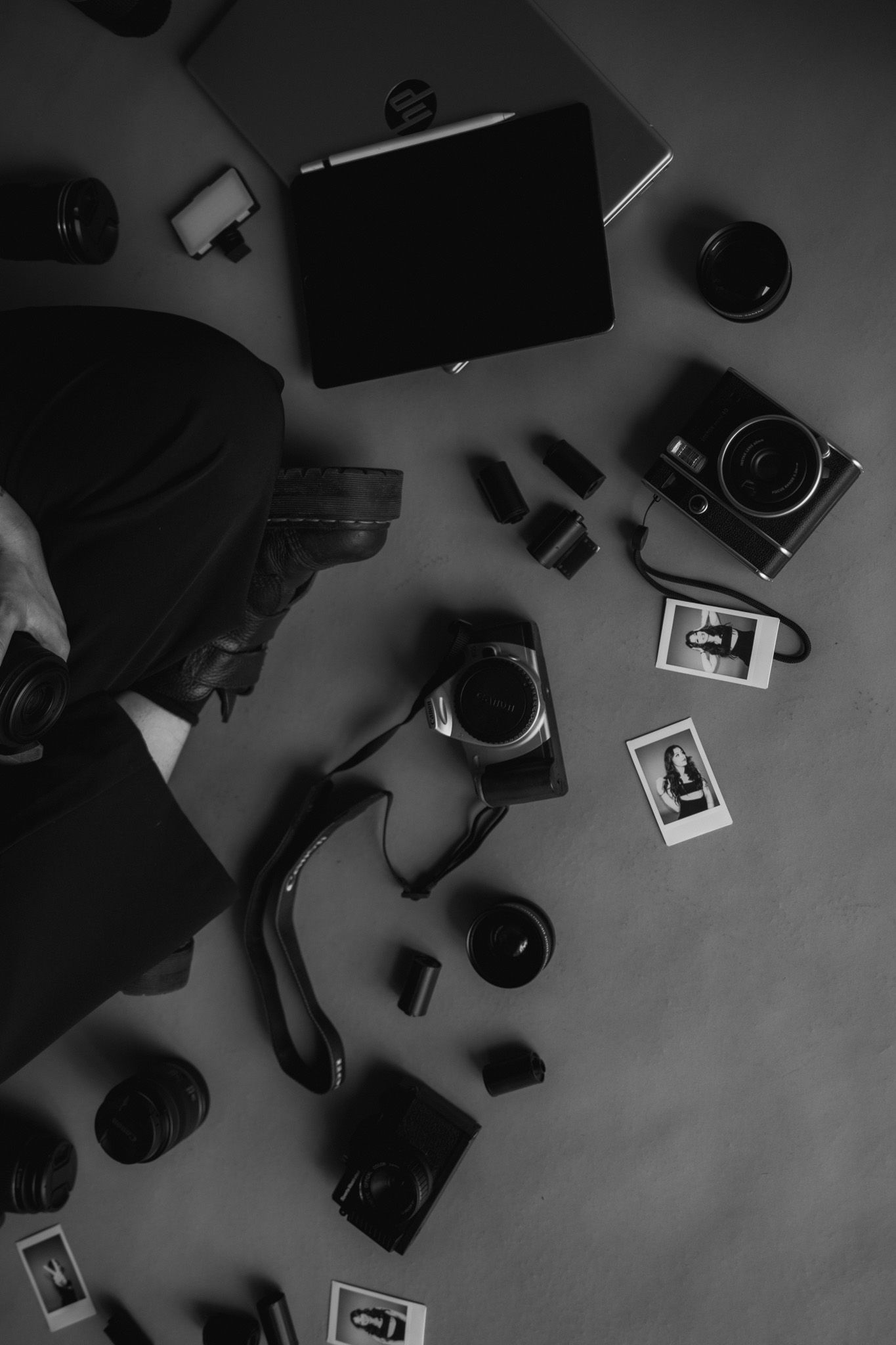 Overhead black and white flatlay of photography equipment including cameras, lenses, and polaroid prints.