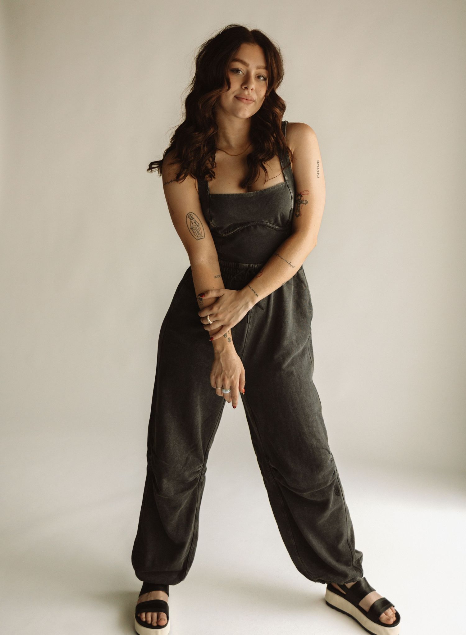 A person poses in a dark jumpsuit against a plain background wearing platform sandals and casual dark attire.