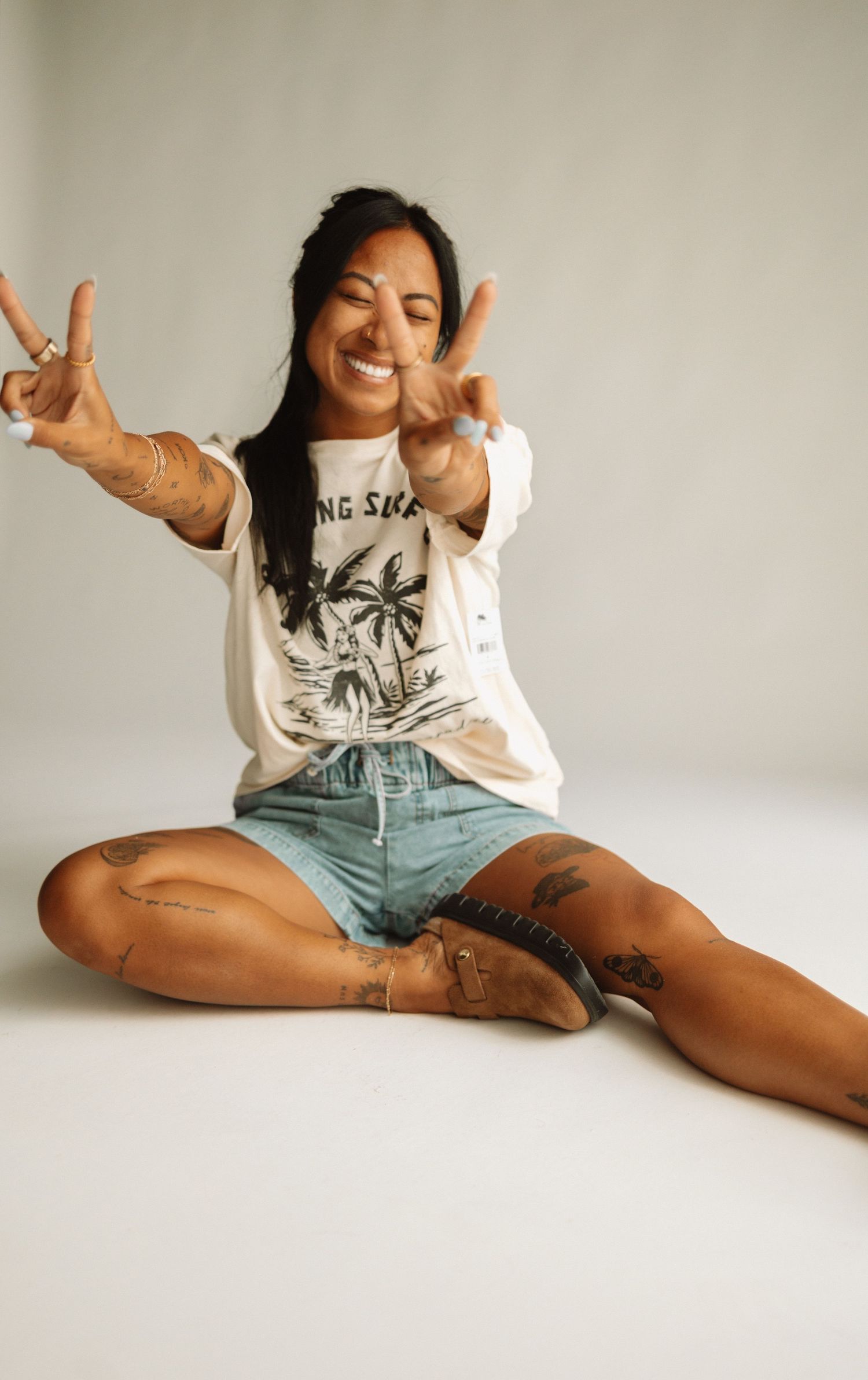Person in casual summer outfit of graphic t-shirt and denim shorts sits cross-legged making peace signs against light background.