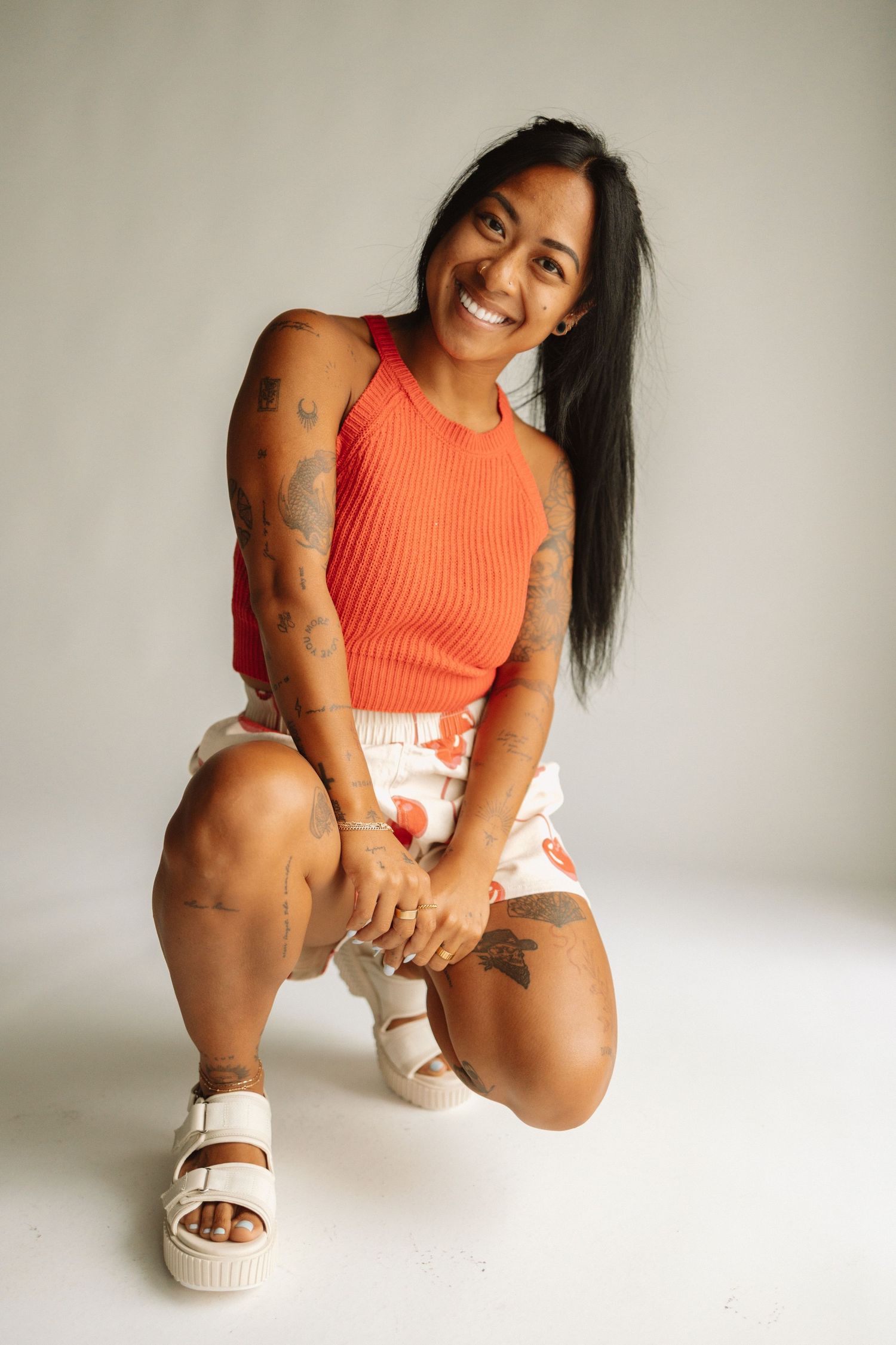 An athletic figure in orange tank top and white shorts poses against white background with tattoos and platform sandals.
