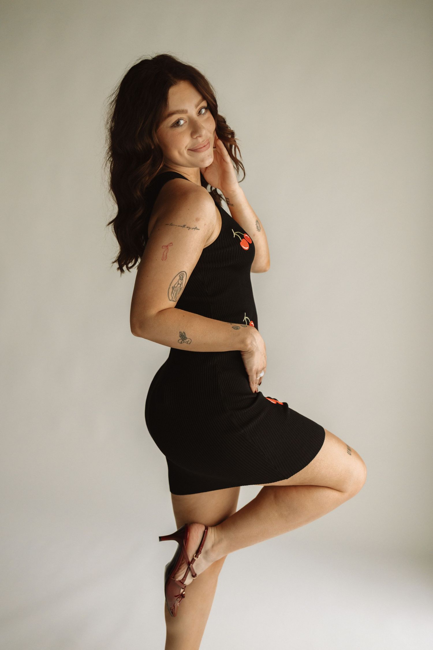 A woman in a black bodycon dress poses against a neutral background showing tattoos on her arms.