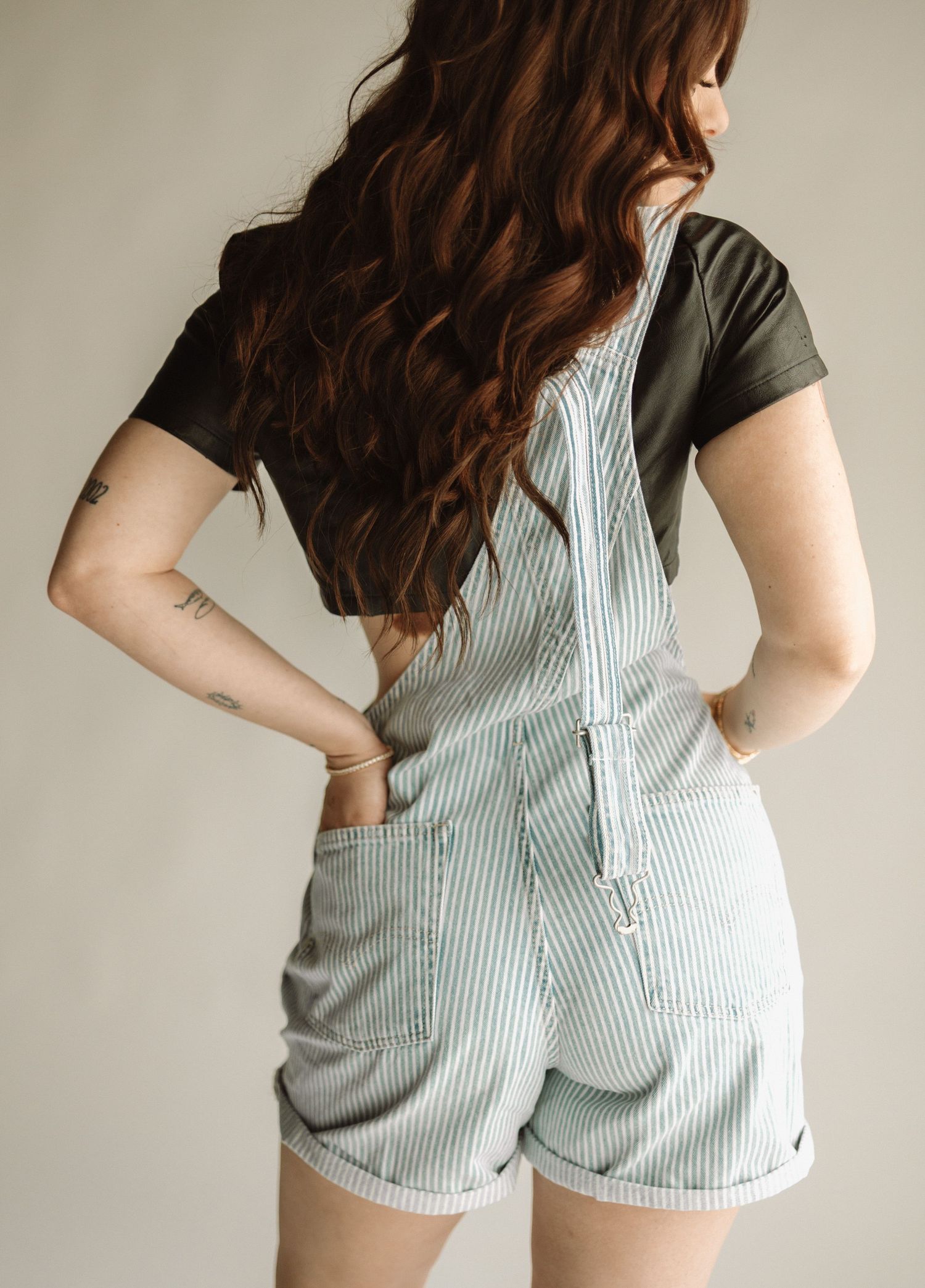 Person wearing striped shortall overalls and black t-shirt shown from behind with long wavy brown hair.