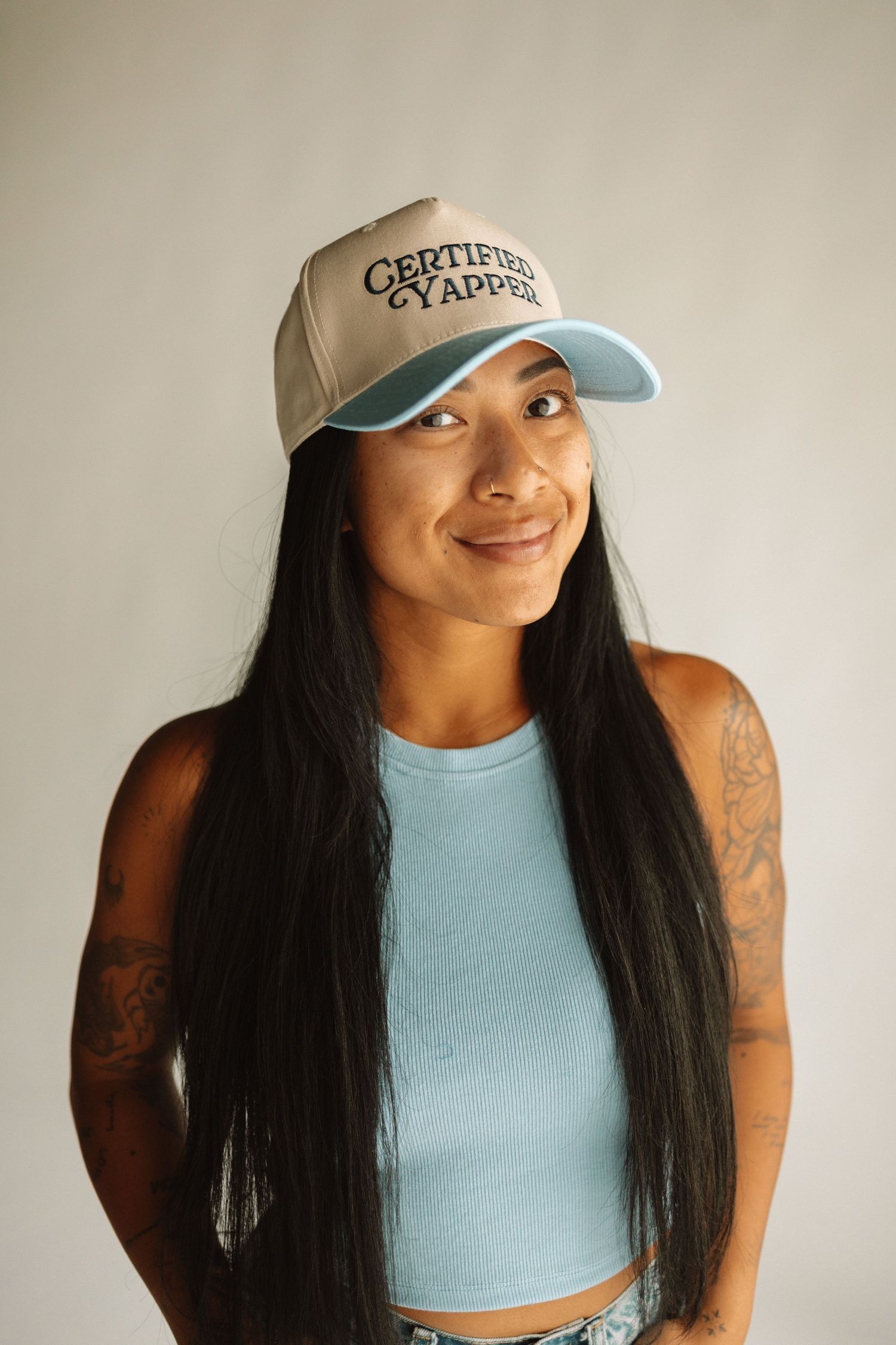Someone wearing a baseball cap and light blue crop top smiles at the camera while showing off arm tattoos and long black hair.
