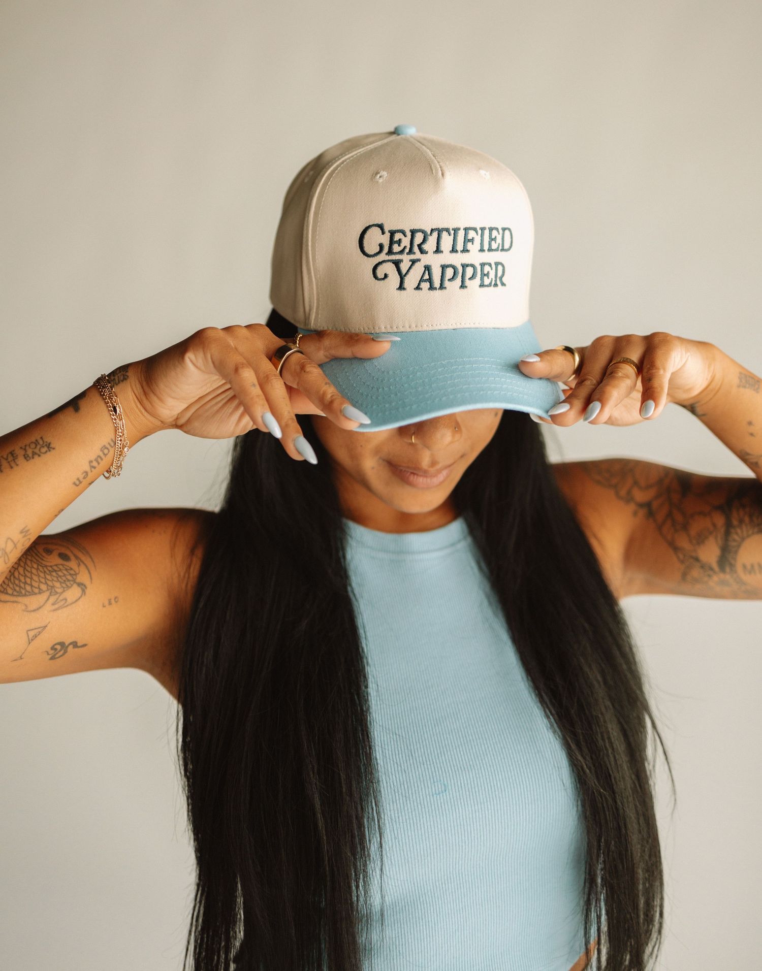 Someone wearing a light blue tank top and baseball cap with 'Certified YAPPer' text adjusts their hat with tattooed arms.