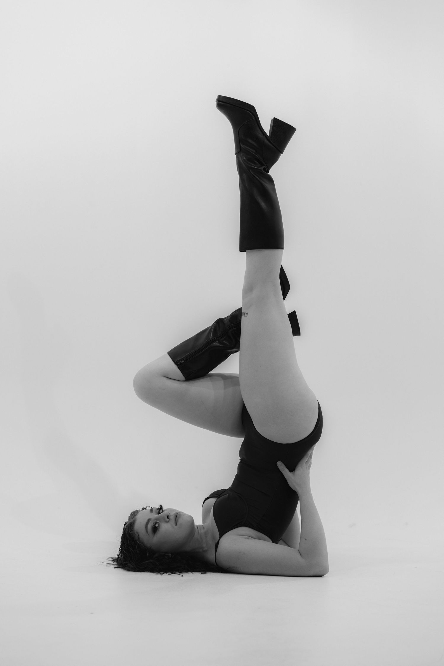 Black and white image of graceful dancer in boots performing upside down pose against white backdrop.