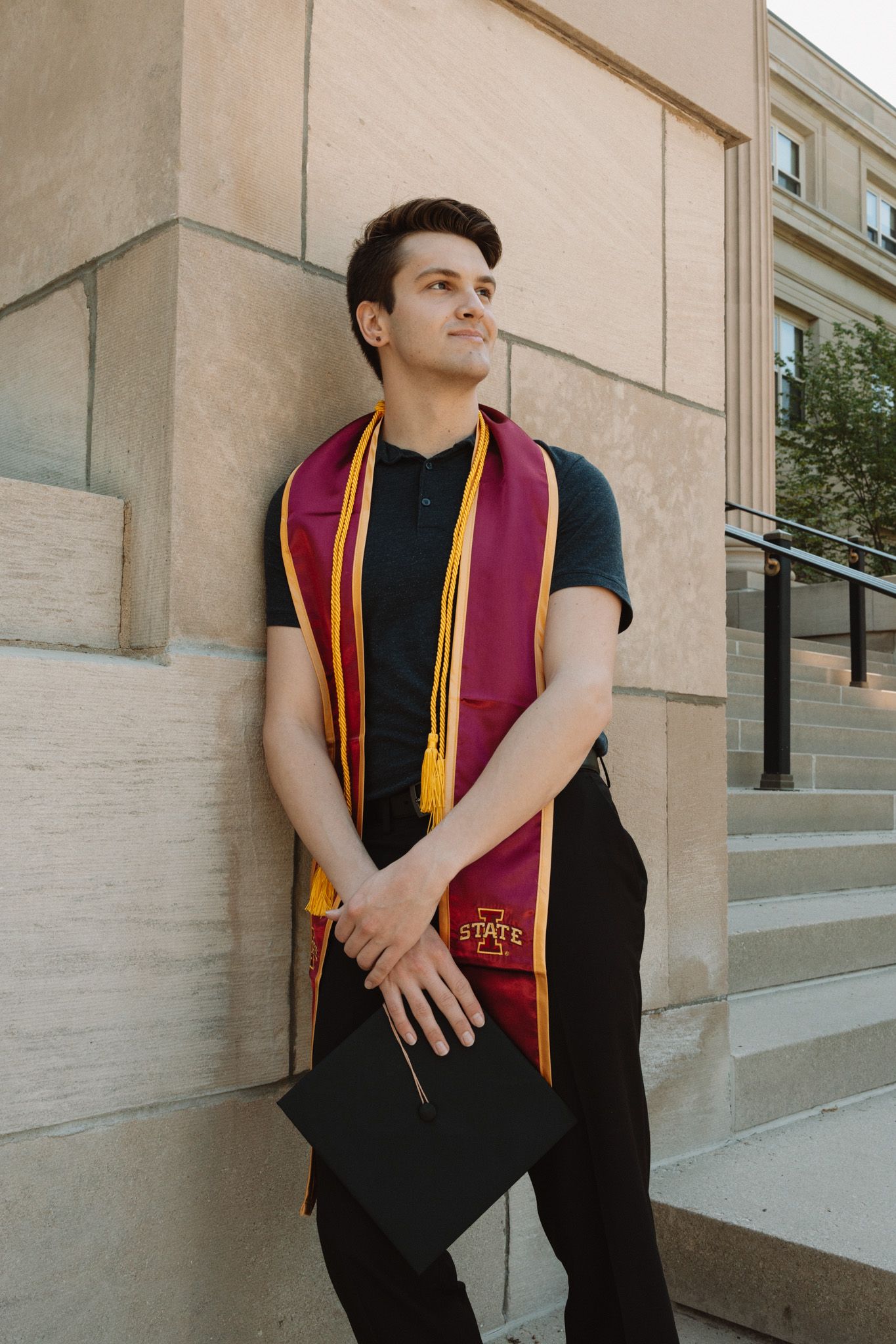 College graduate wearing maroon and gold graduation stole with black attire poses on campus steps.