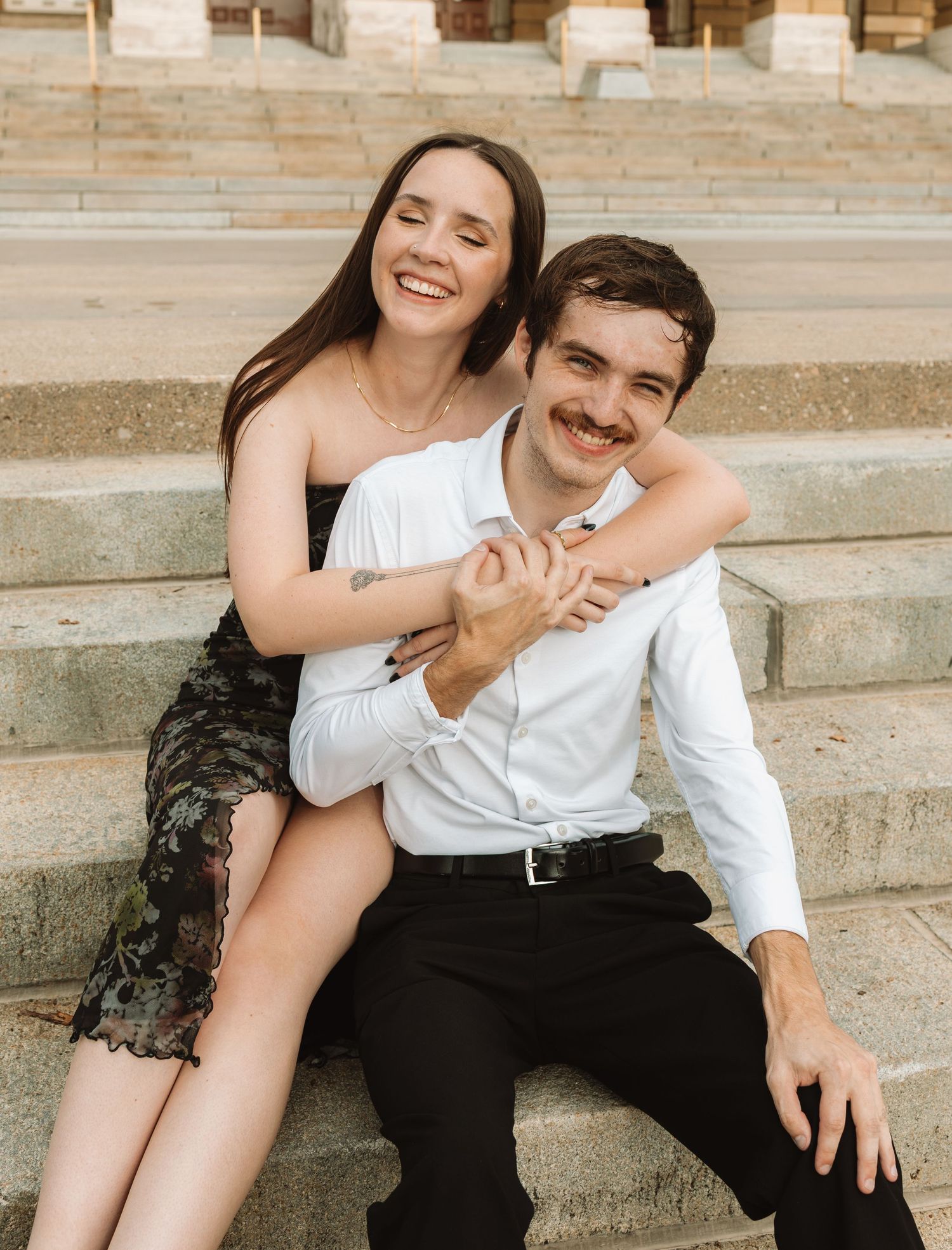 A couple shares a joyful moment sitting together on outdoor stone steps in casual attire.