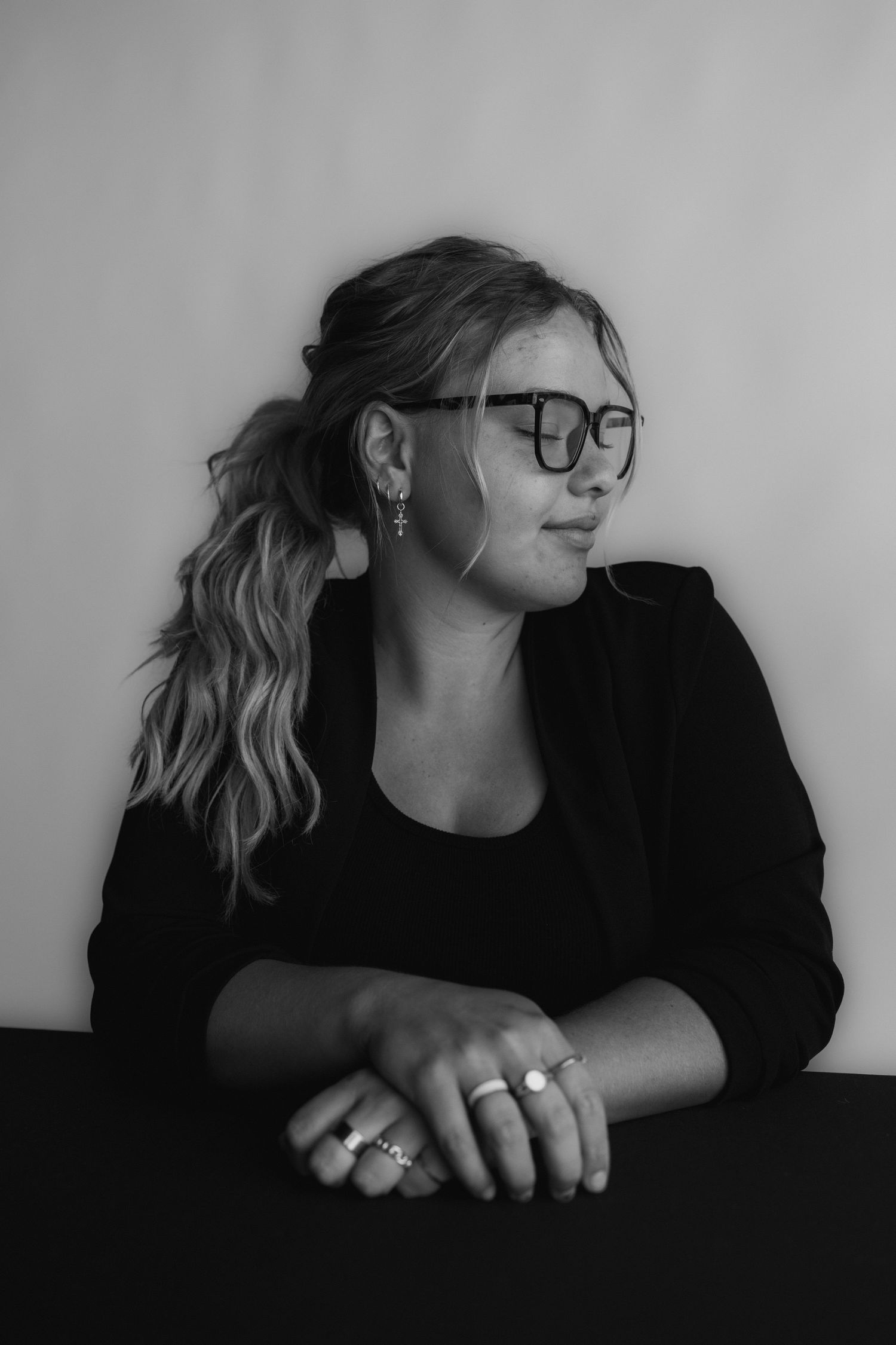 Black and white portrait of someone in glasses and a dark blazer looking to the side while seated at a table.