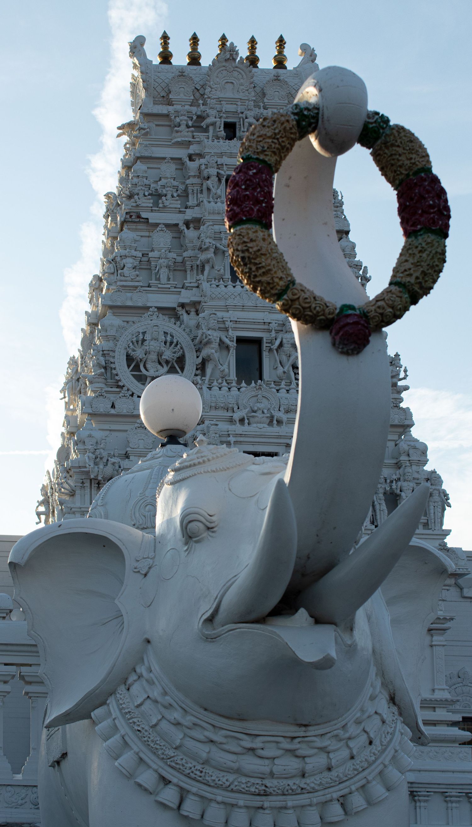 White elephant statue holds a decorative wreath in front of an ornate Hindu temple tower with detailed carvings.
