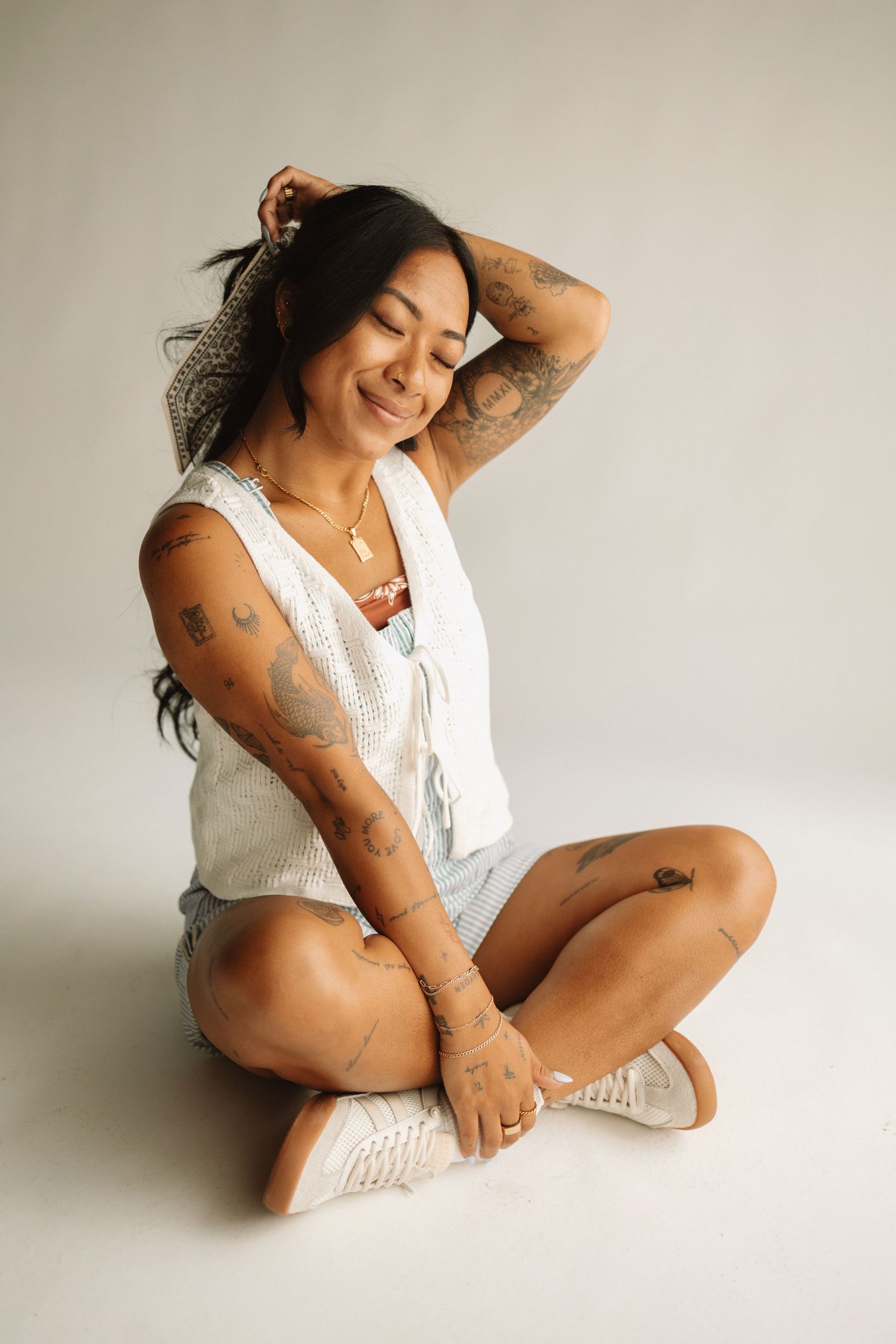 Carefree young woman in white tank top and sneakers sits cross-legged on floor, showing off arm tattoos while smiling.