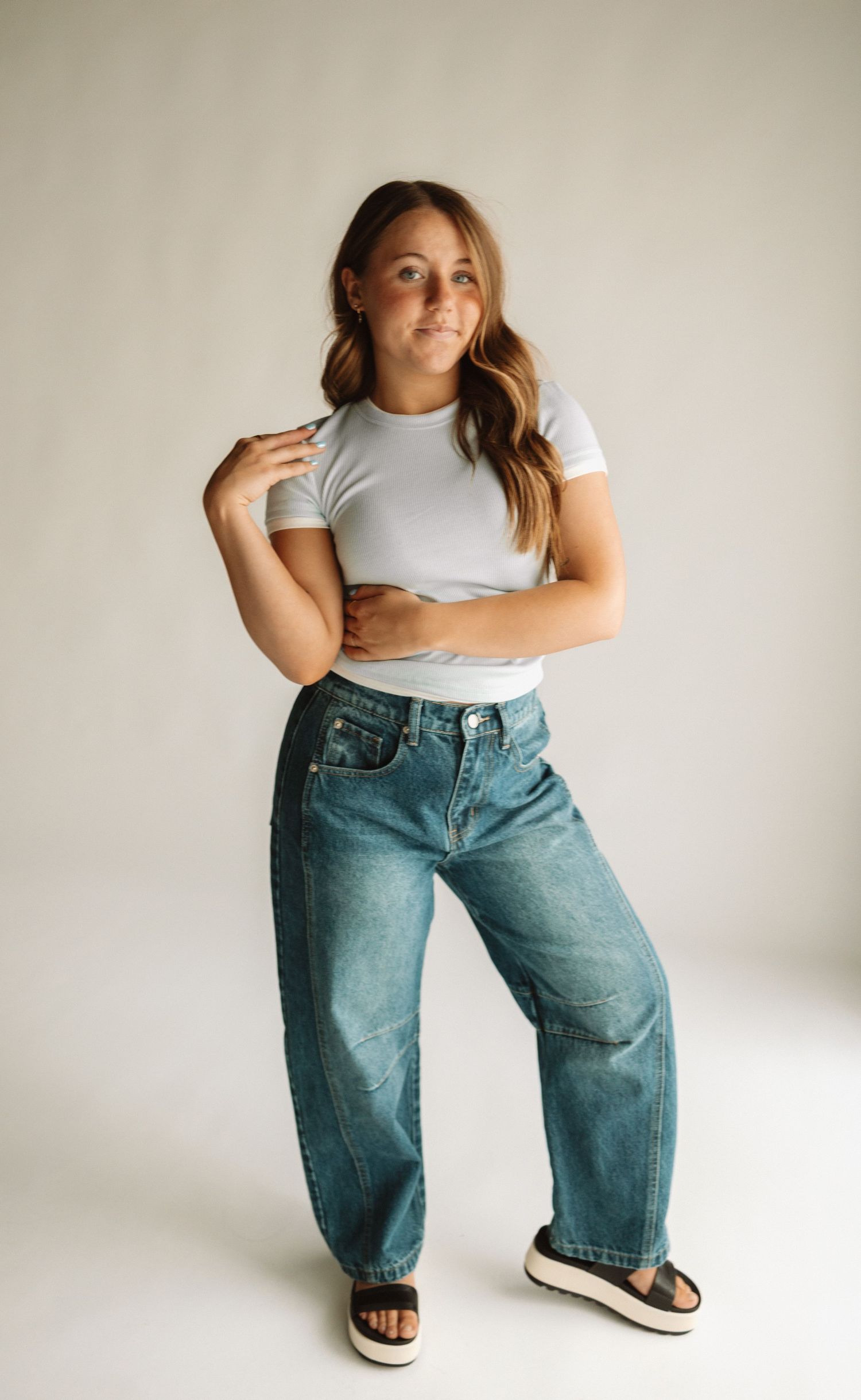 Fashion model wearing a casual grey t-shirt and baggy blue jeans poses in front of a plain white wall.
