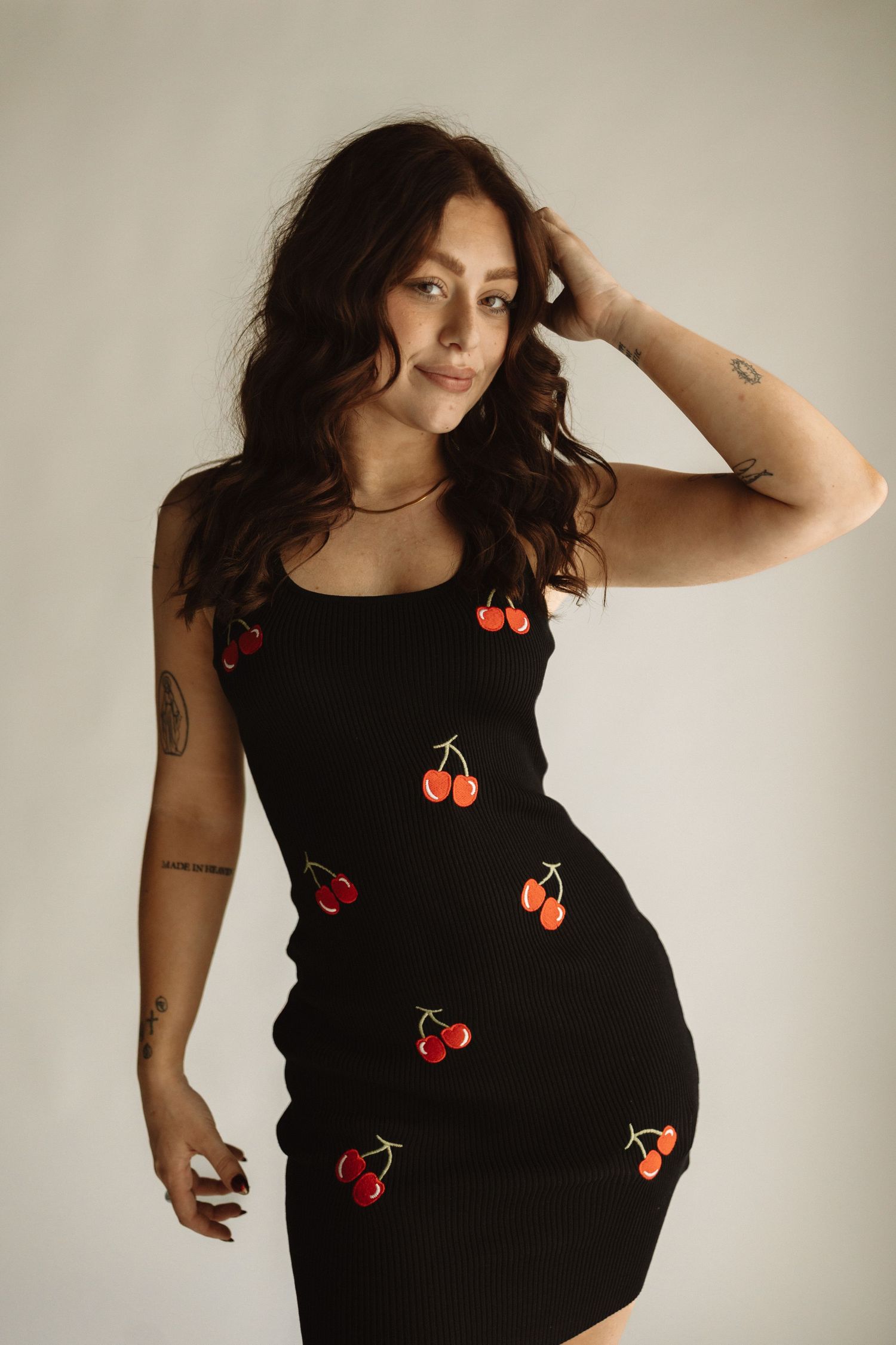 Person in black dress with red cherry appliques poses against neutral background in vintage-inspired fashion photoshoot series.