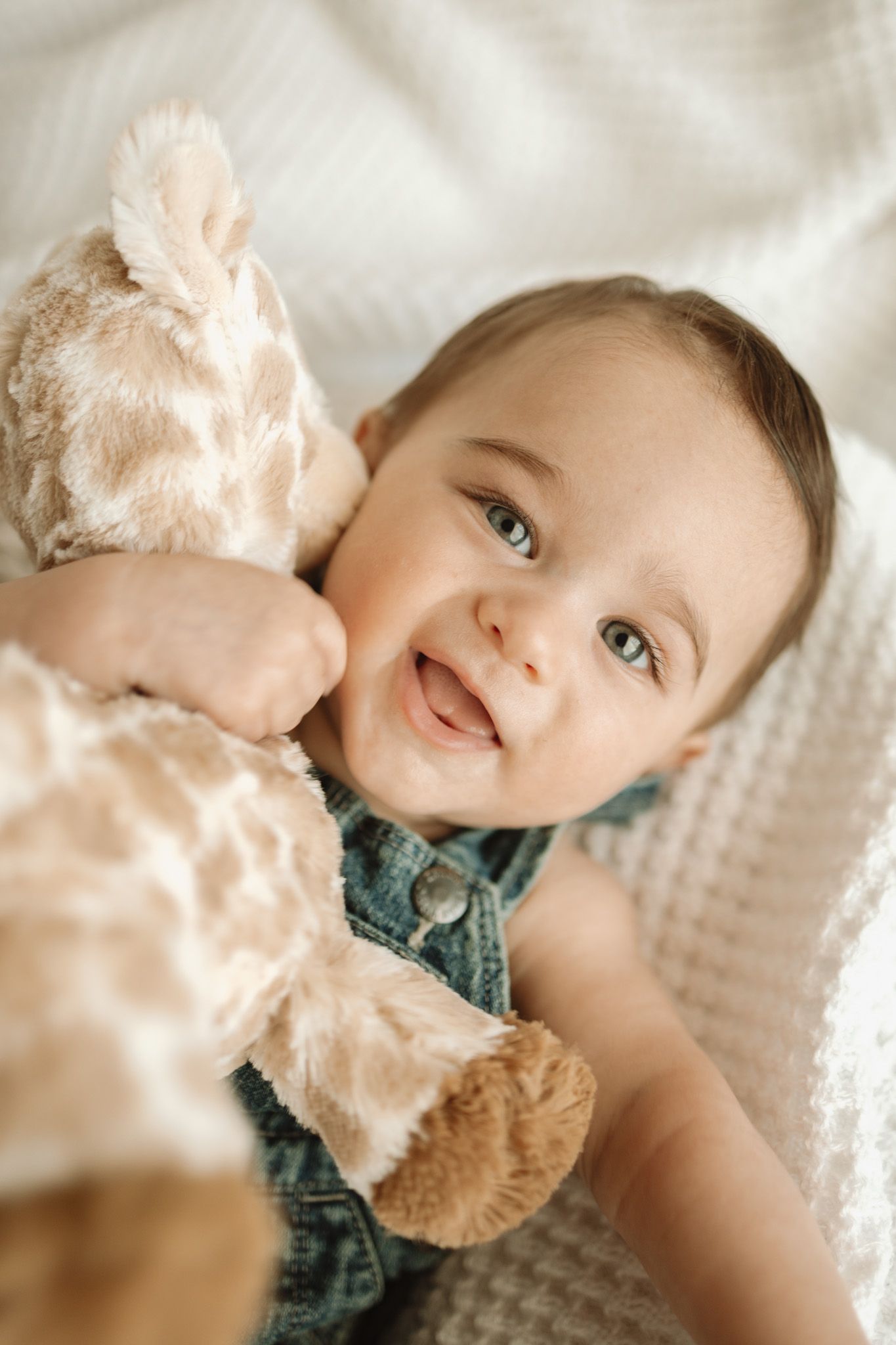 A smiling baby in denim clothing hugs a soft plush stuffed animal while lying on a textured white blanket.