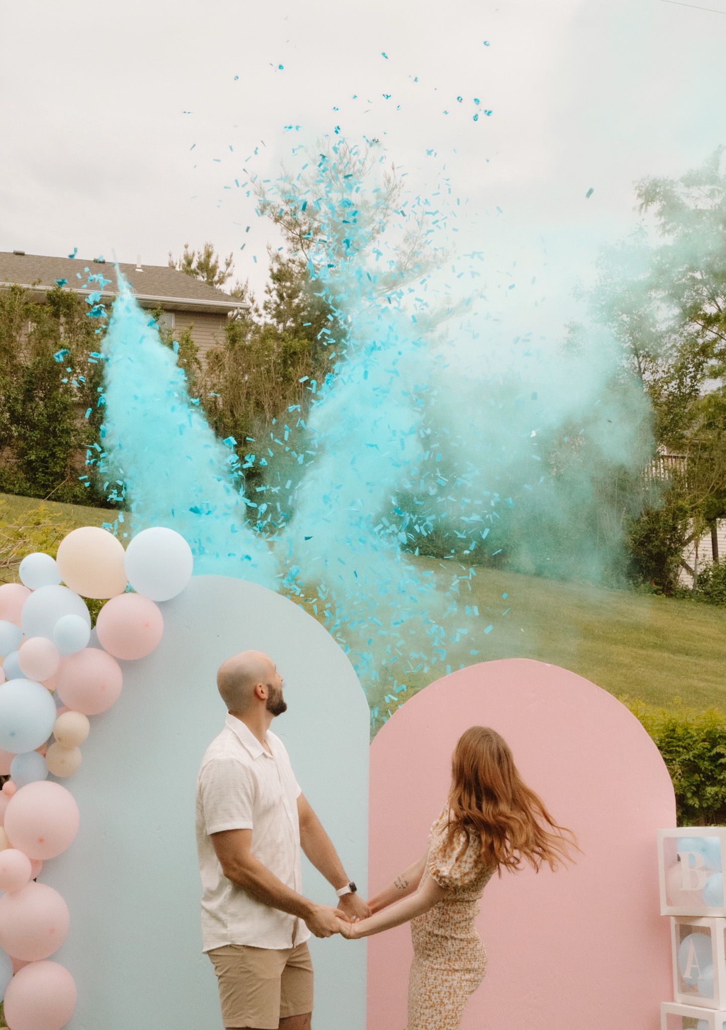 Gender reveal party celebration with blue powder explosion and pink and white balloon arch in an outdoor garden setting.