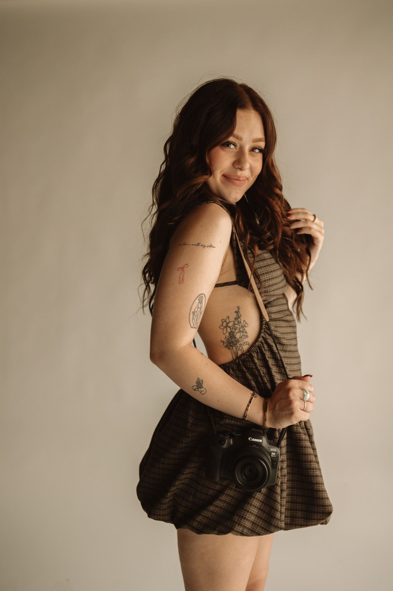 Photographer in striped brown dress with visible tattoos poses with vintage camera against beige wall in natural light.