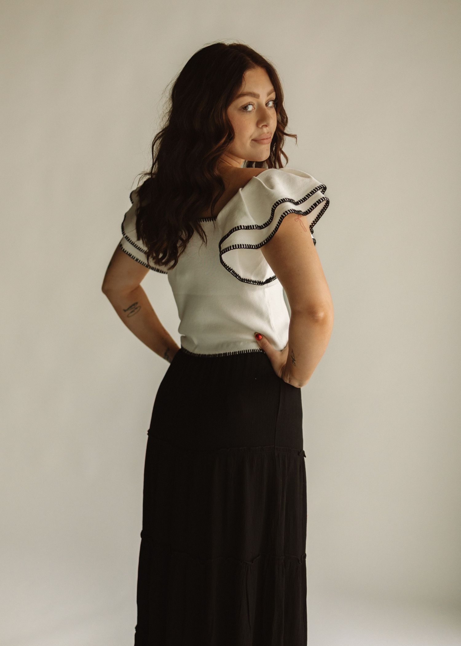 A person poses against neutral wall wearing white ruffled blouse and black maxi skirt with hands in pockets.