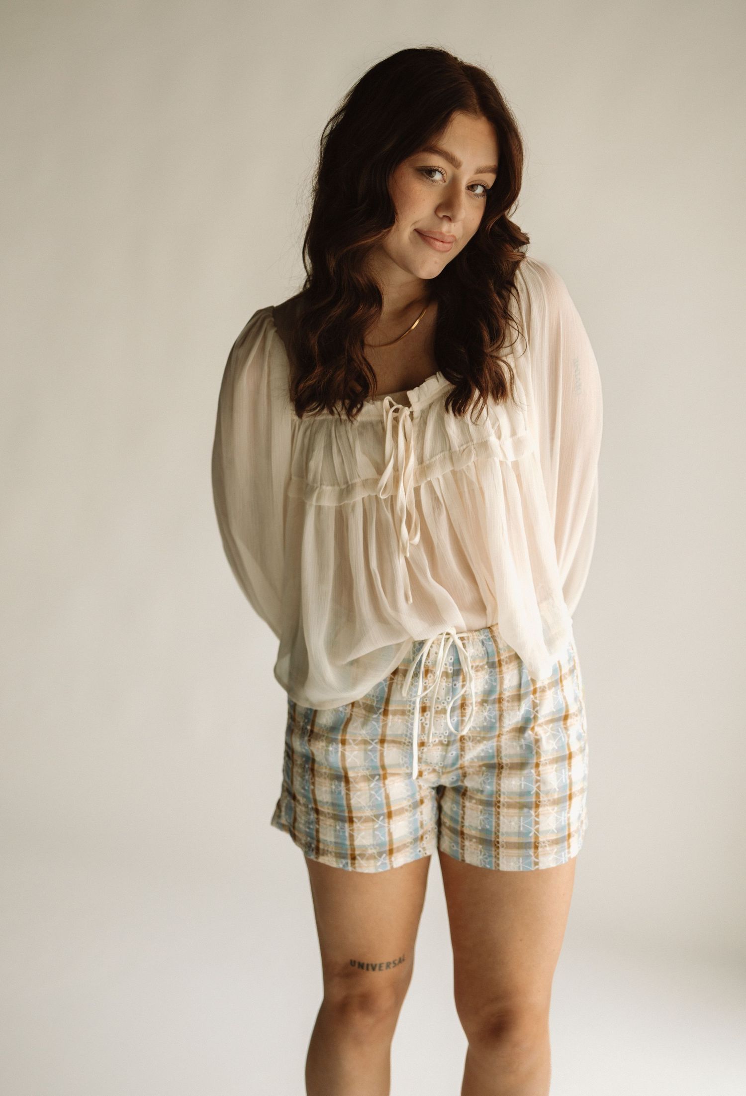 Person in a flowy cream blouse and plaid shorts poses against a neutral wall in a minimalist fashion photo shoot.