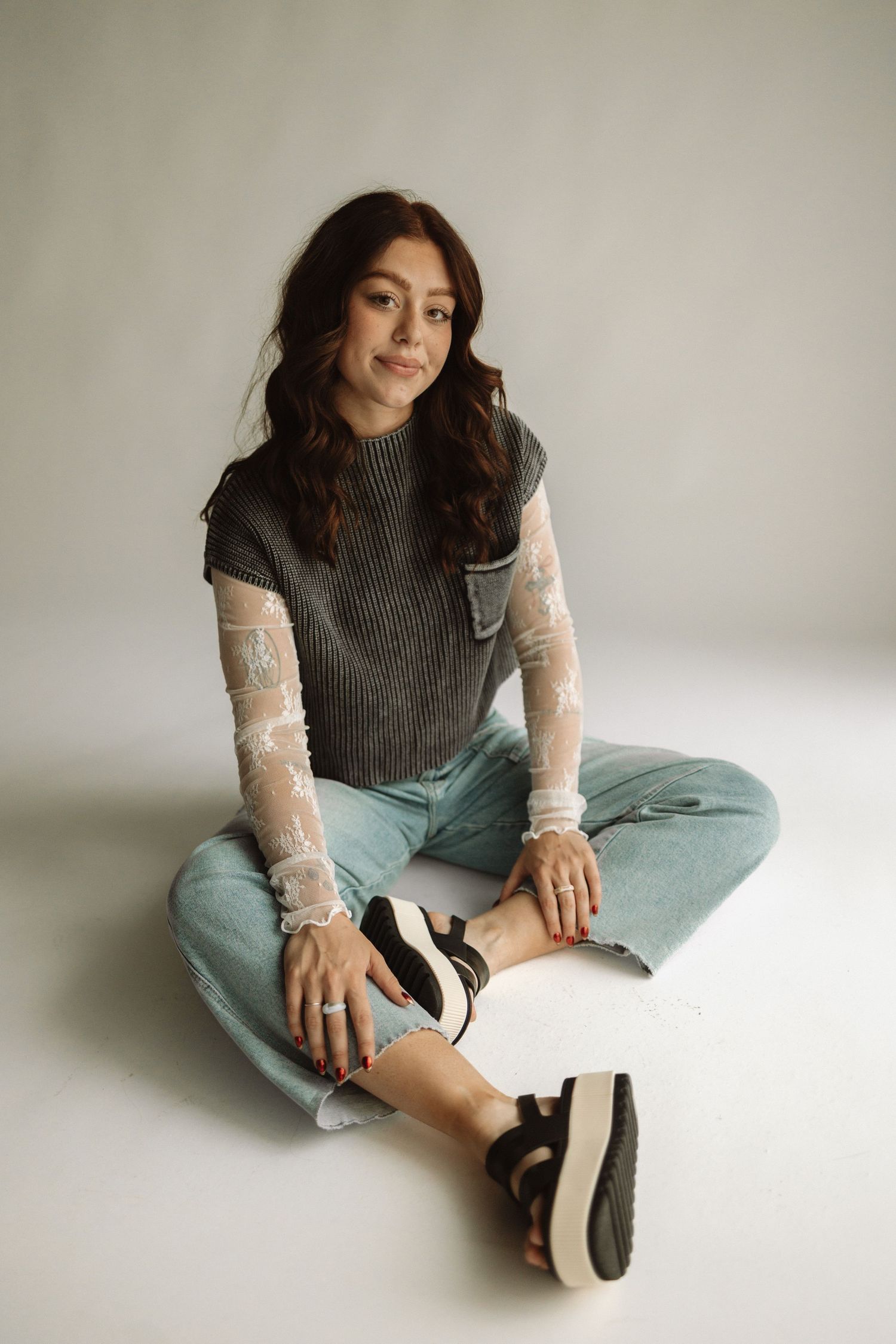Casual portrait of someone in a dark sleeveless sweater, light jeans and platform sandals sitting cross-legged on white floor.