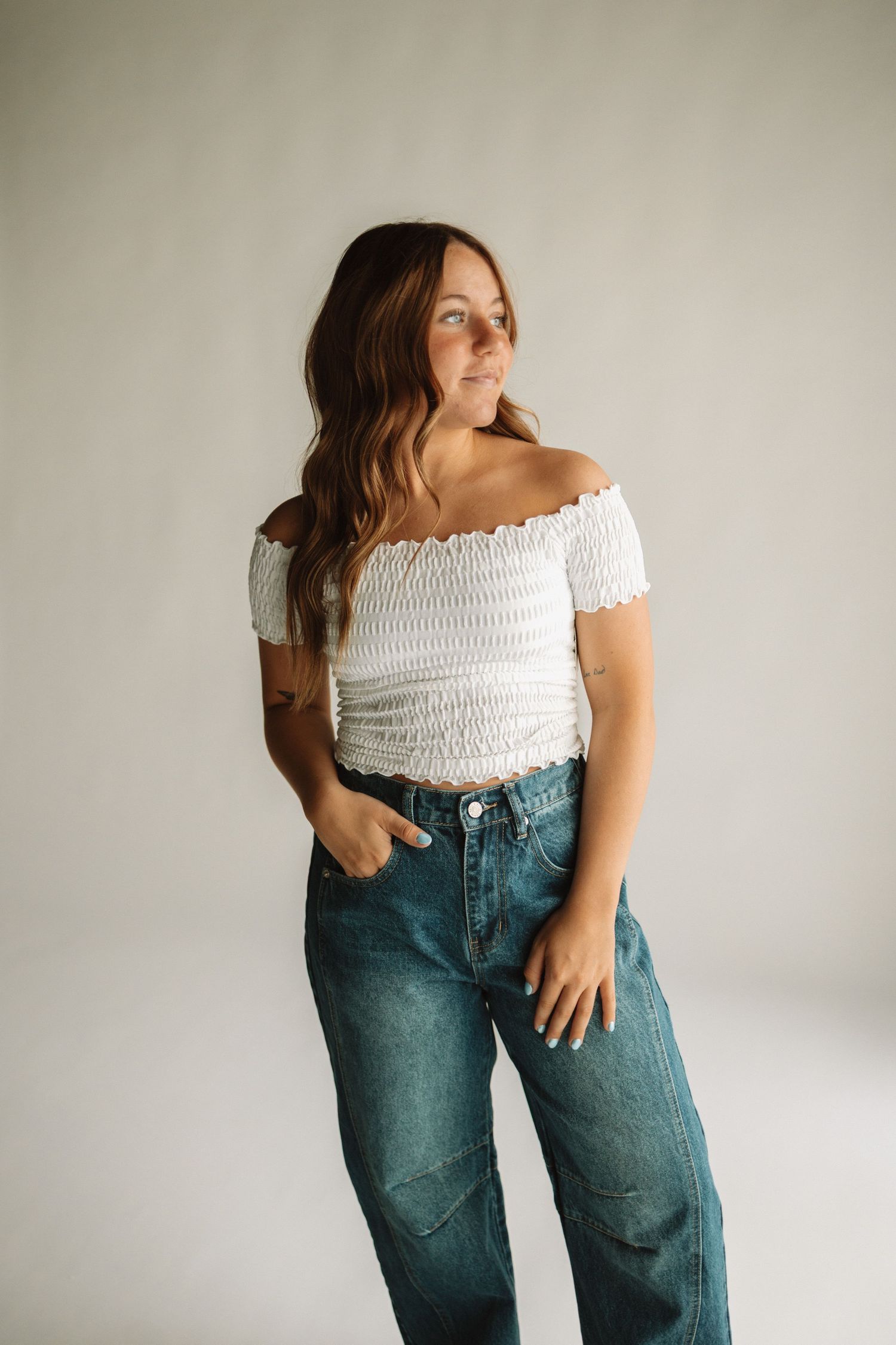 Fashion model wearing white smocked off-shoulder crop top with relaxed blue jeans against neutral backdrop.