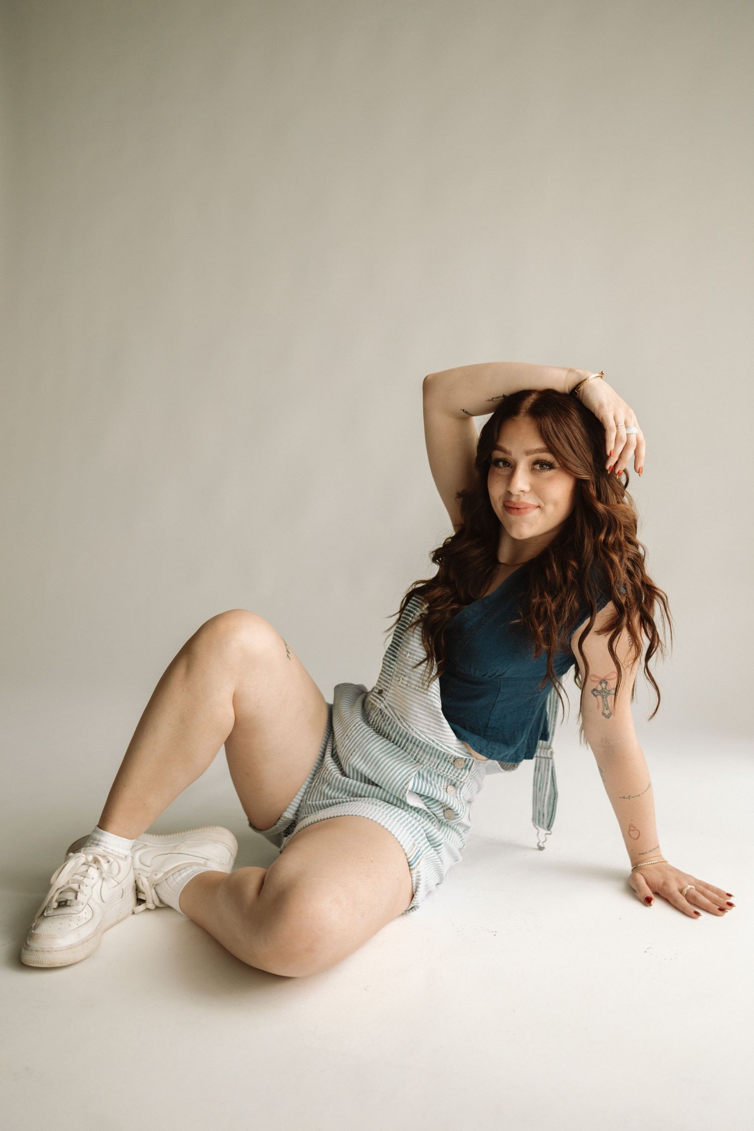 Person in casual attire with long wavy hair poses while sitting on white backdrop wearing sneakers and shorts.