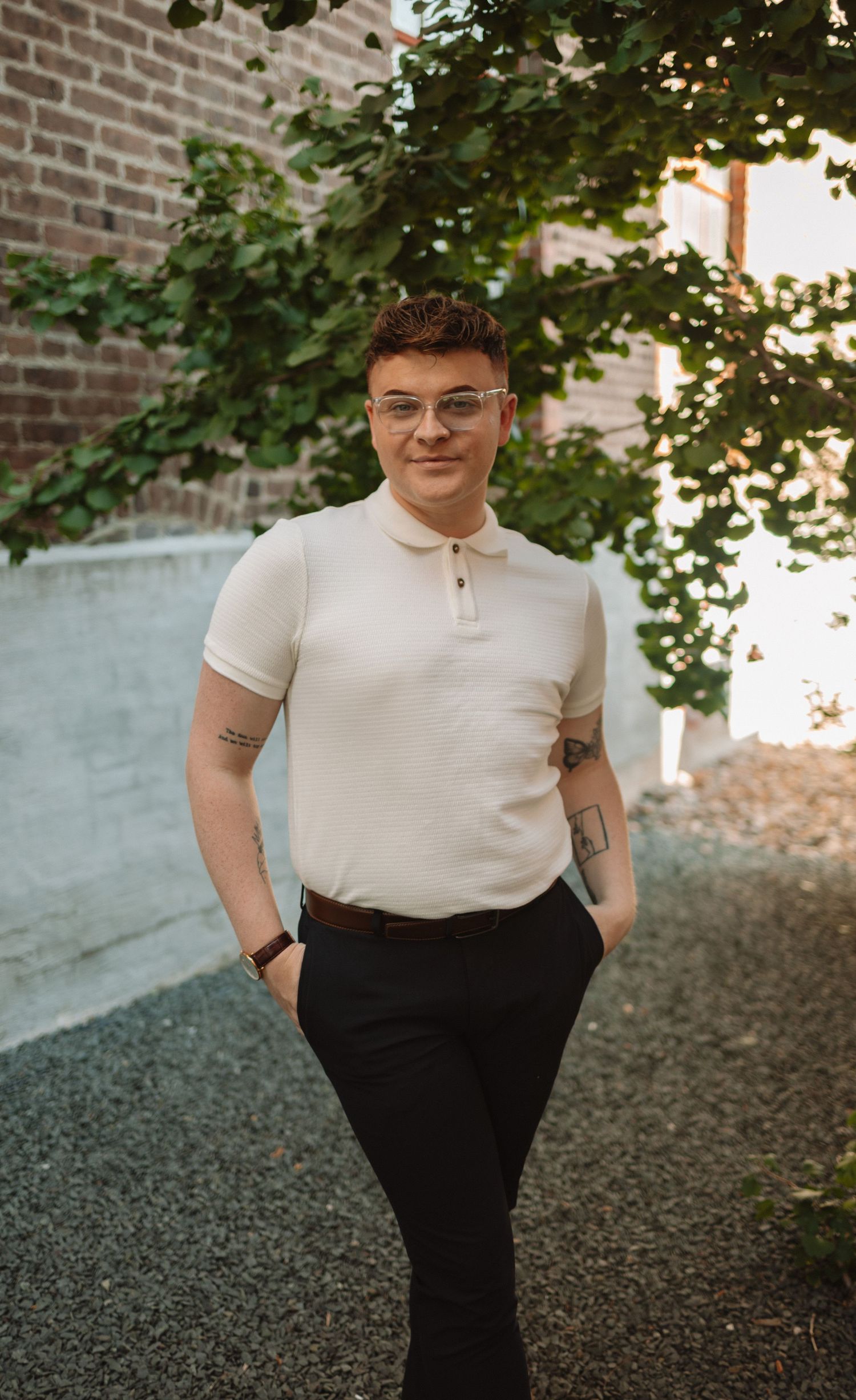A person in a cream polo shirt and dark pants stands casually against a brick wall with green foliage in the background.