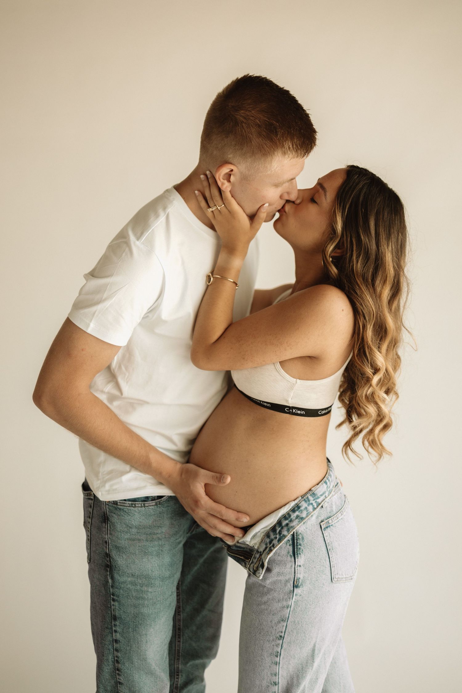 A romantic maternity photoshoot featuring couple in jeans and white shirt against neutral background.