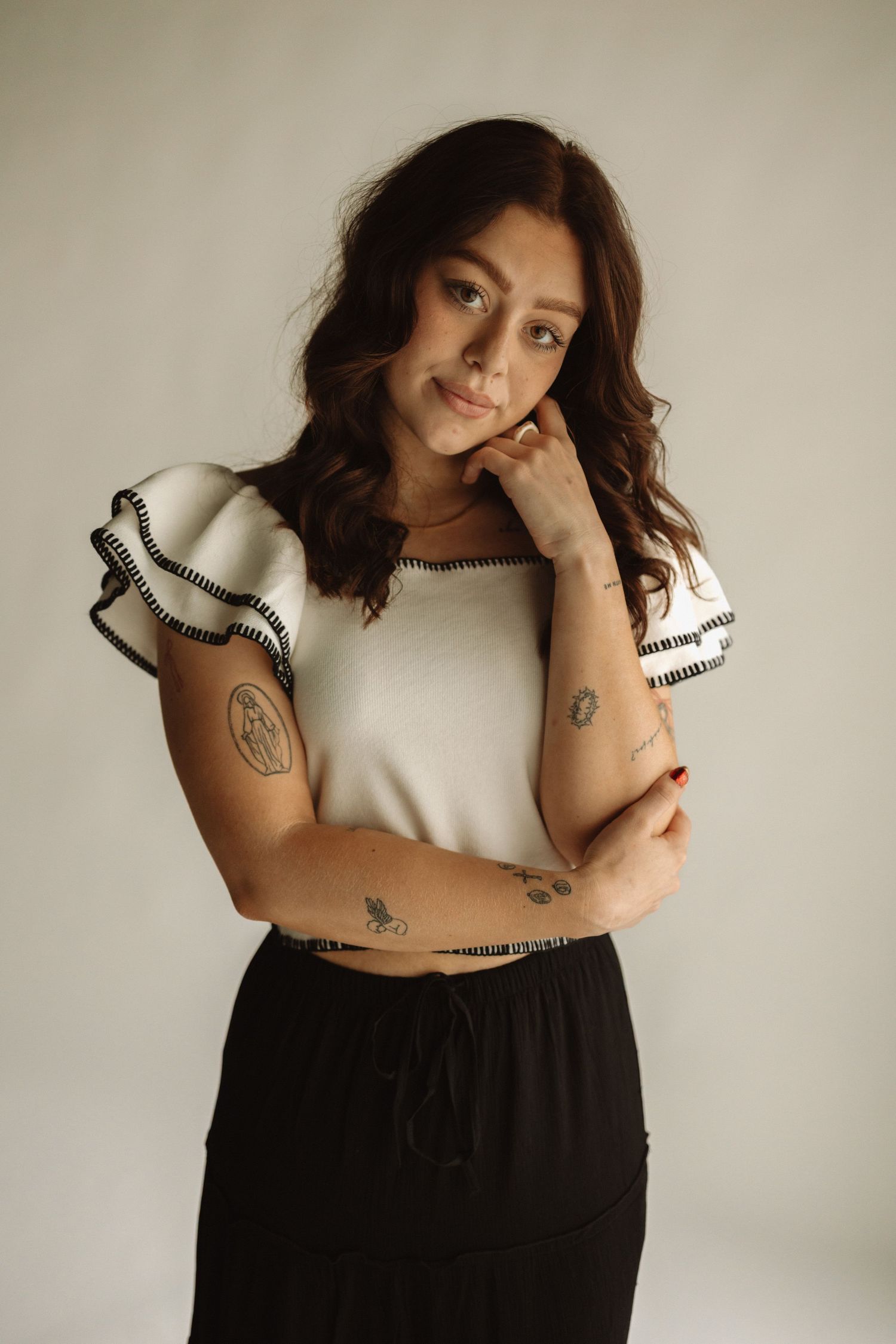 A person poses in a black and cream outfit with ruffled sleeves and visible tattoos against a light background.