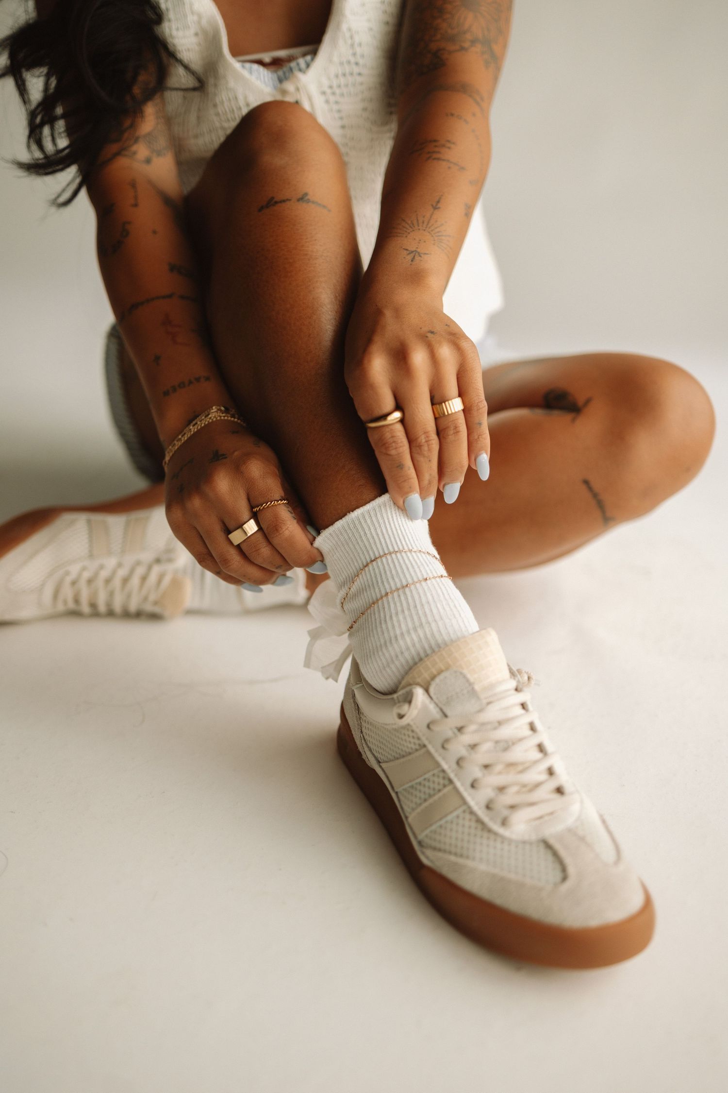 Person wearing white ribbed socks and retro-style white sneakers with gum soles sits on white surface.