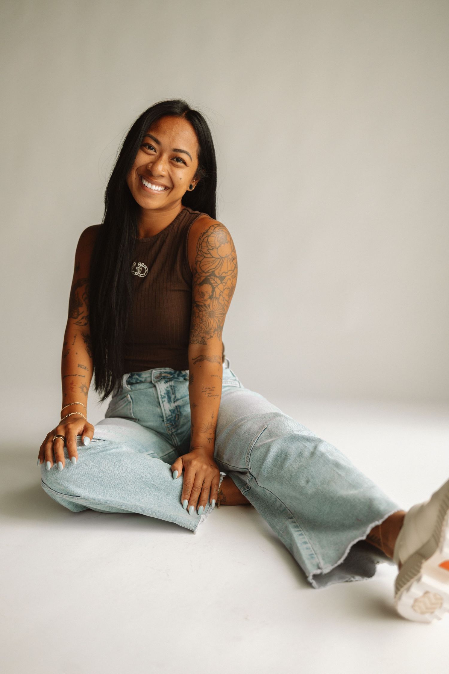 A person smiles while sitting on the floor wearing a brown tank top, light wash baggy jeans and sneakers.