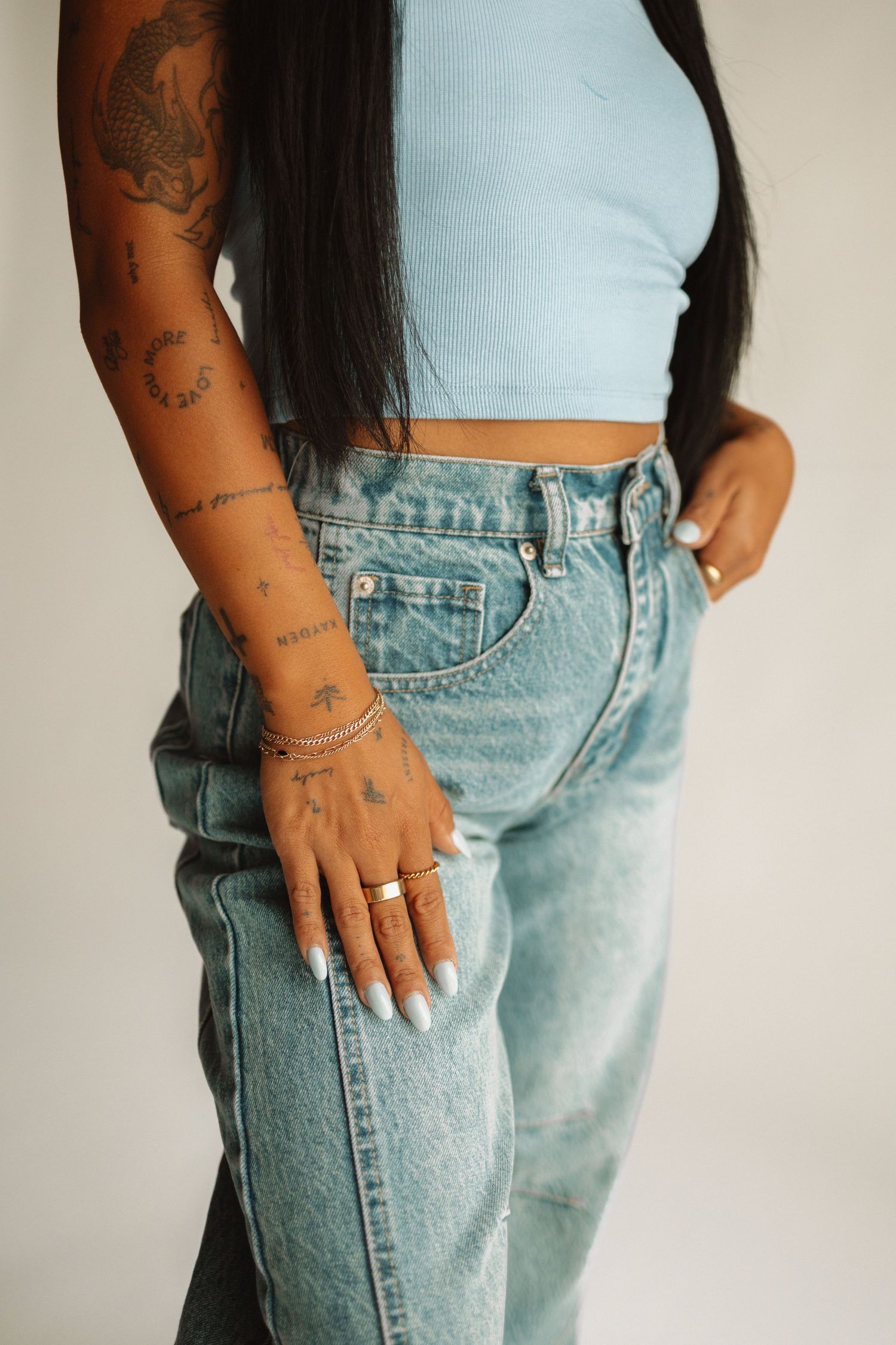 Cropped view of someone wearing light wash baggy jeans and white crop top with visible arm tattoos and rings.
