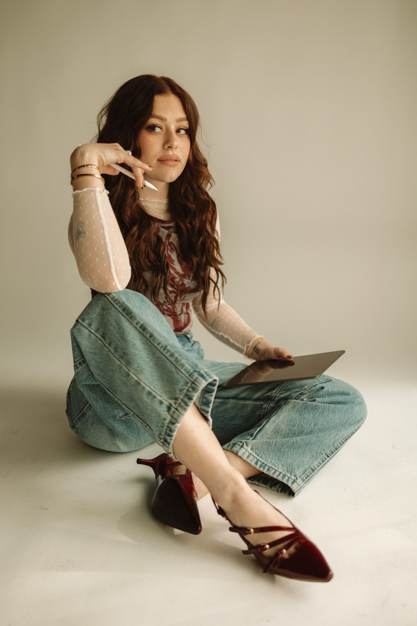 A person in a turtleneck sweater and wide-leg jeans sits on the floor wearing burgundy flats against a neutral background.