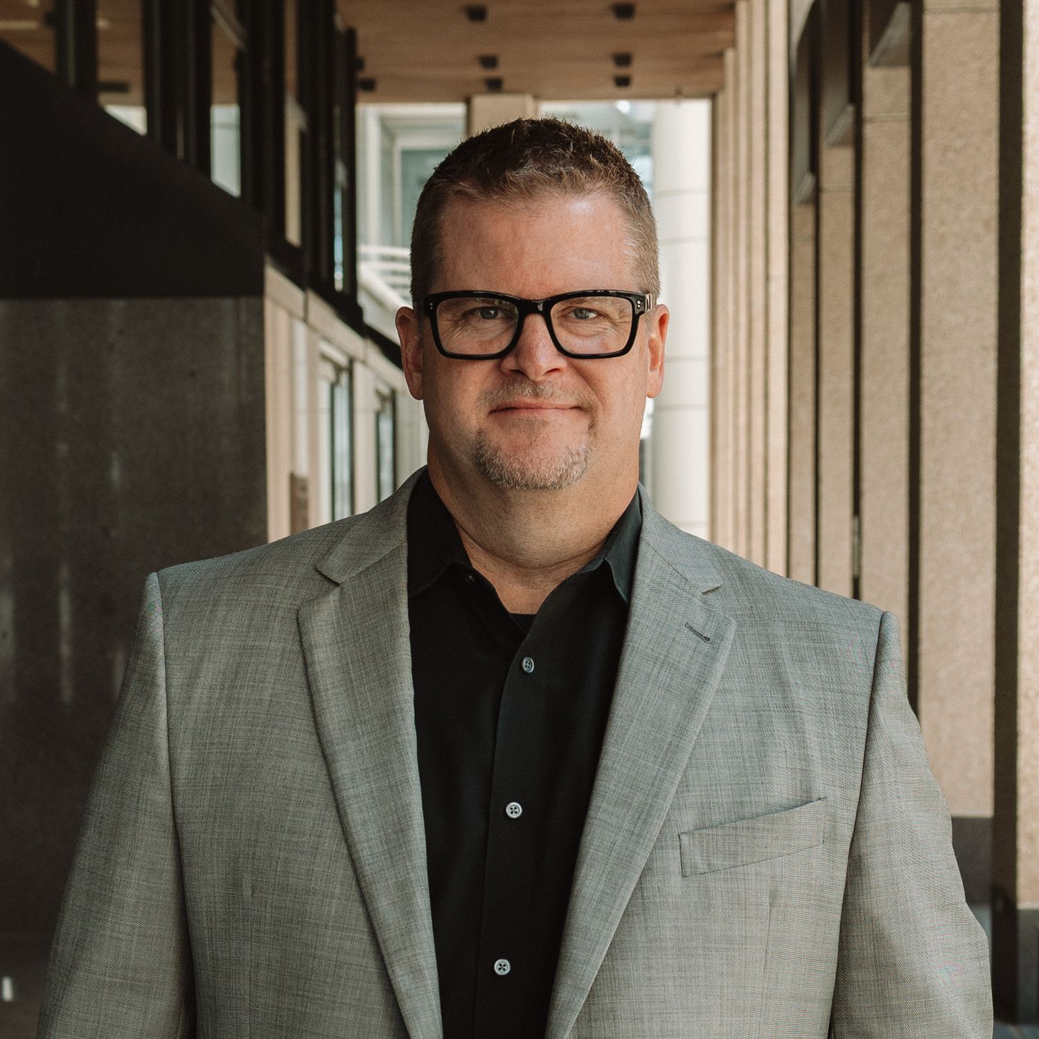 A person in a gray suit jacket and black shirt stands against a beige background in a formal setting.
