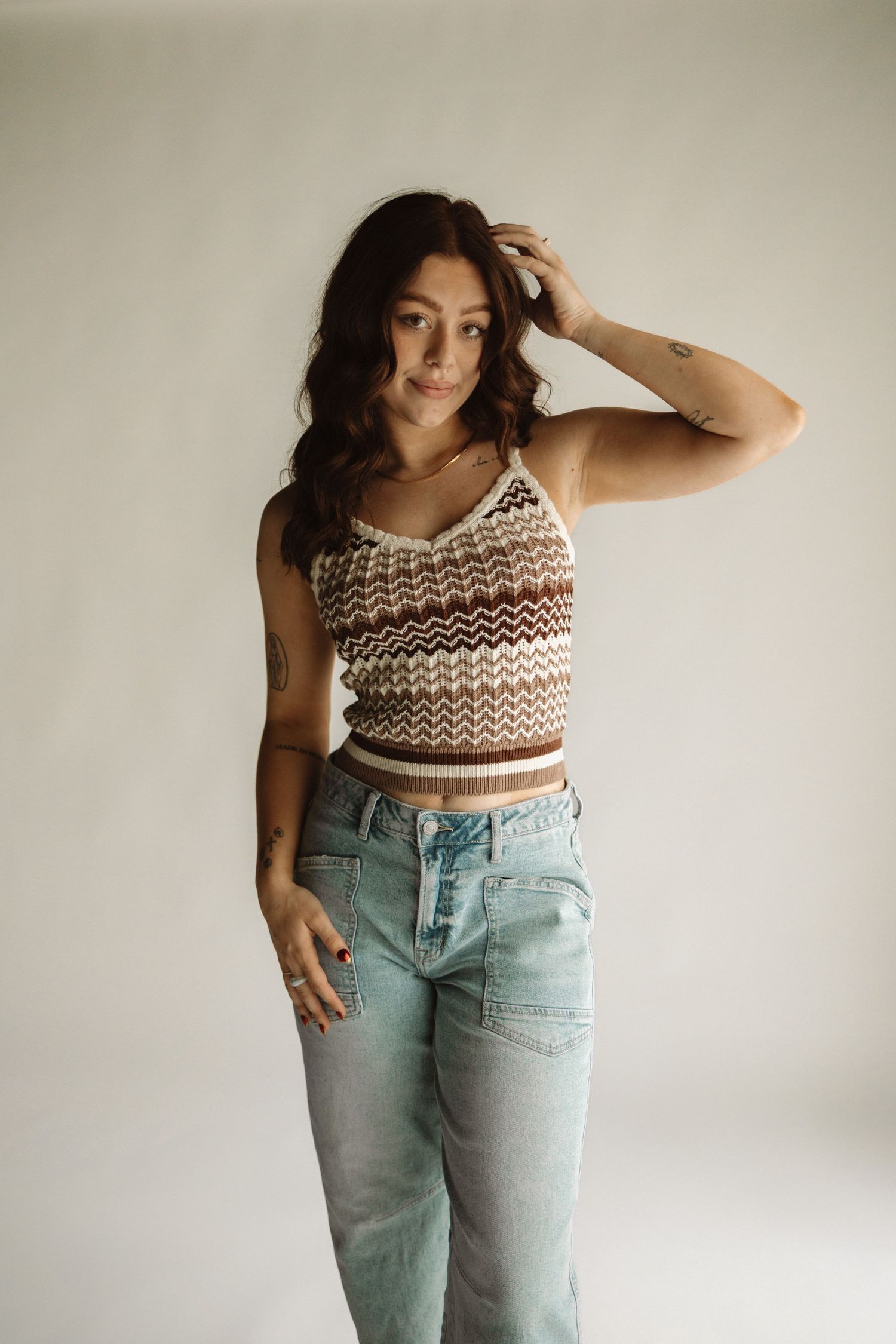 Model poses in brown and white knit crop top and light wash jeans against neutral wall background.