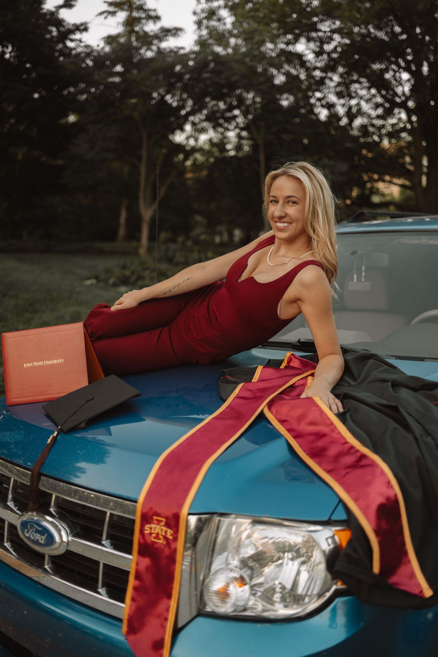 A person in a burgundy dress and gold sash poses on the hood of a blue car at sunset.
