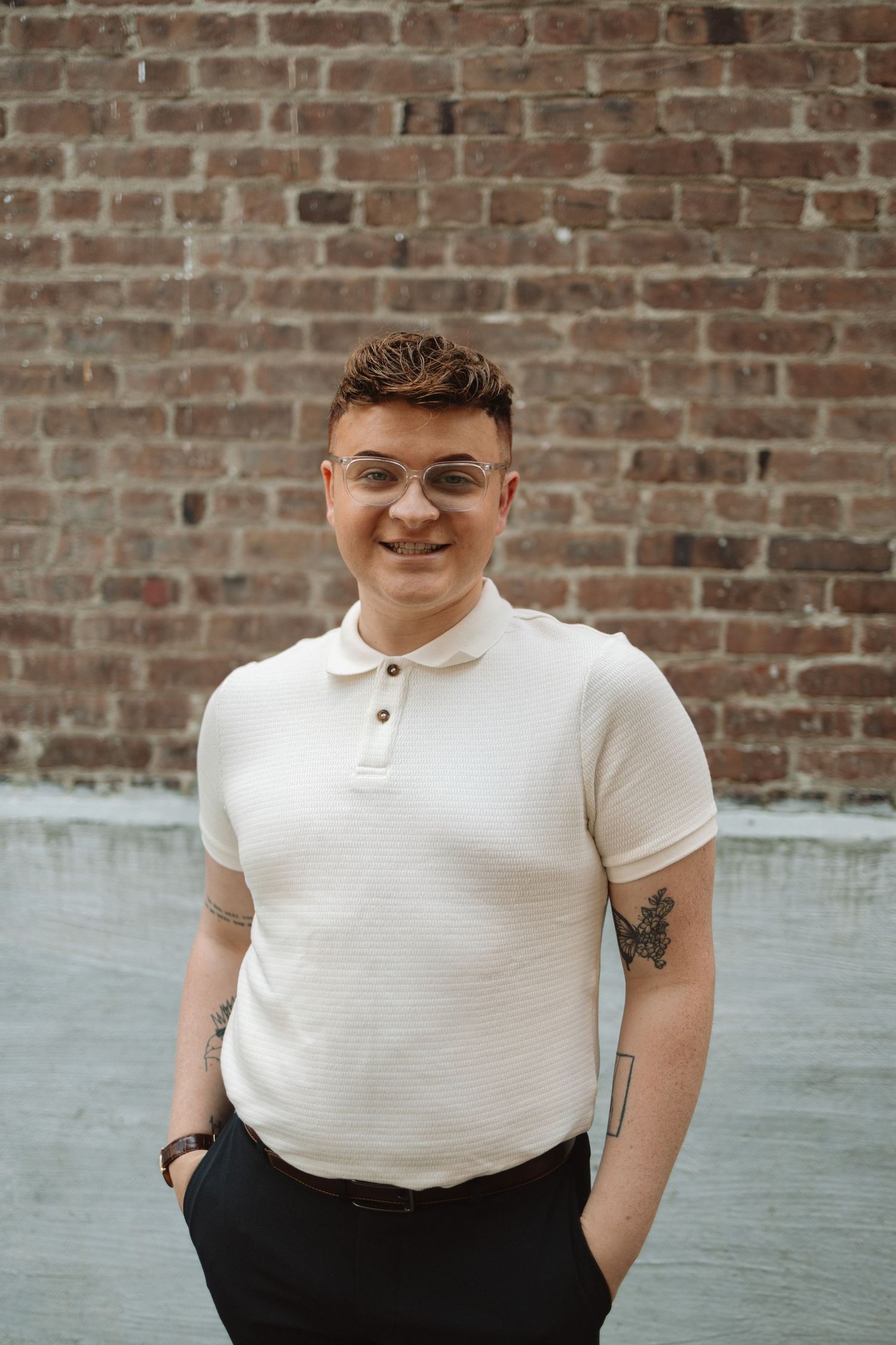 Person wearing a white polo shirt and dark pants poses casually against a rustic brick wall background.
