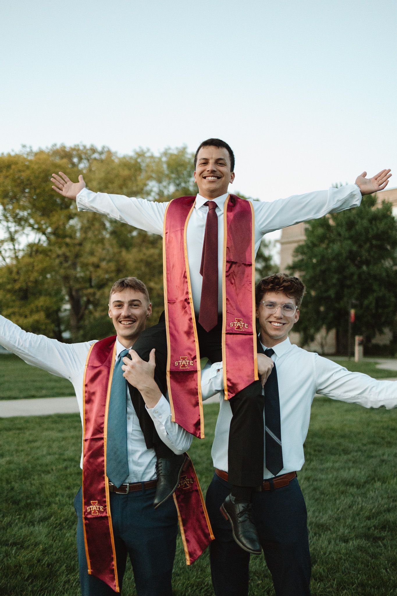 Graduates in white shirts and burgundy stoles celebrate joyfully on a grassy lawn.