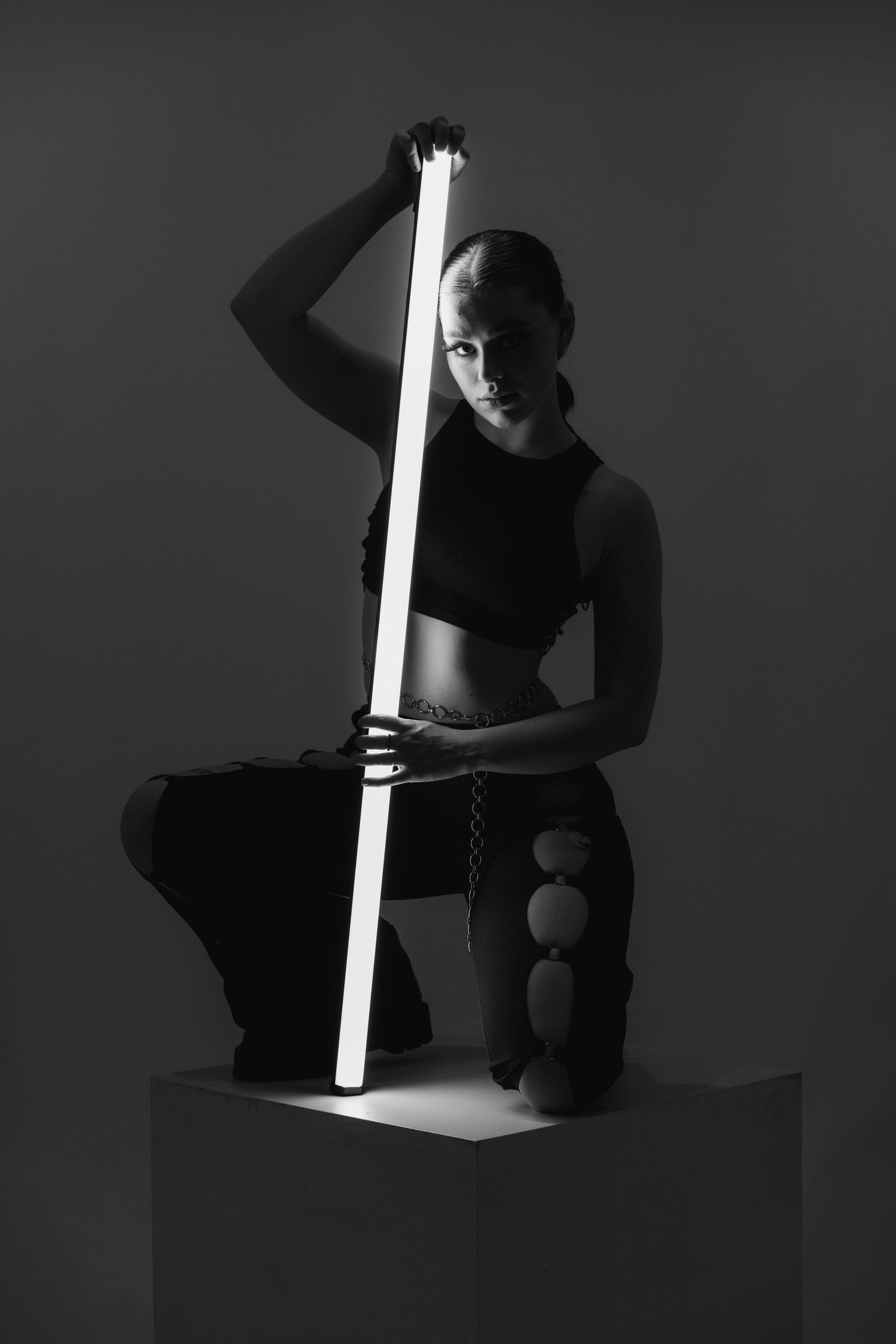 A dramatic black and white artistic portrait with a person holding a glowing light rod while sitting on a pedestal.