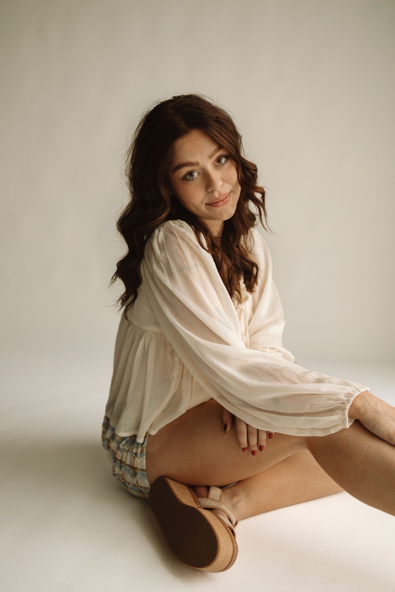 Portrait of someone seated in a white flowy blouse and distressed shorts against a neutral backdrop.