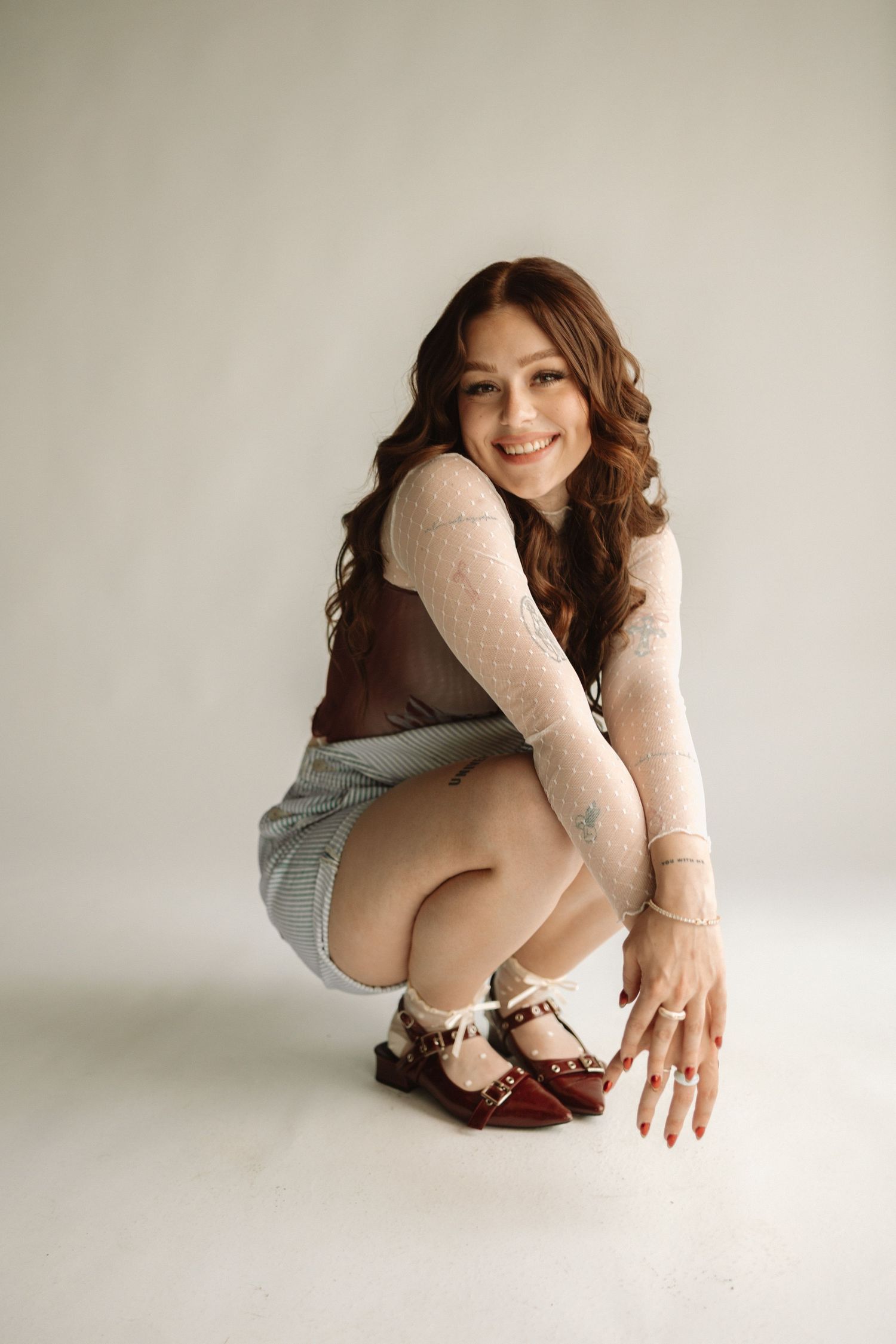 A woman in a cream sweater and denim shorts crouches while wearing brown oxford shoes against a white background.