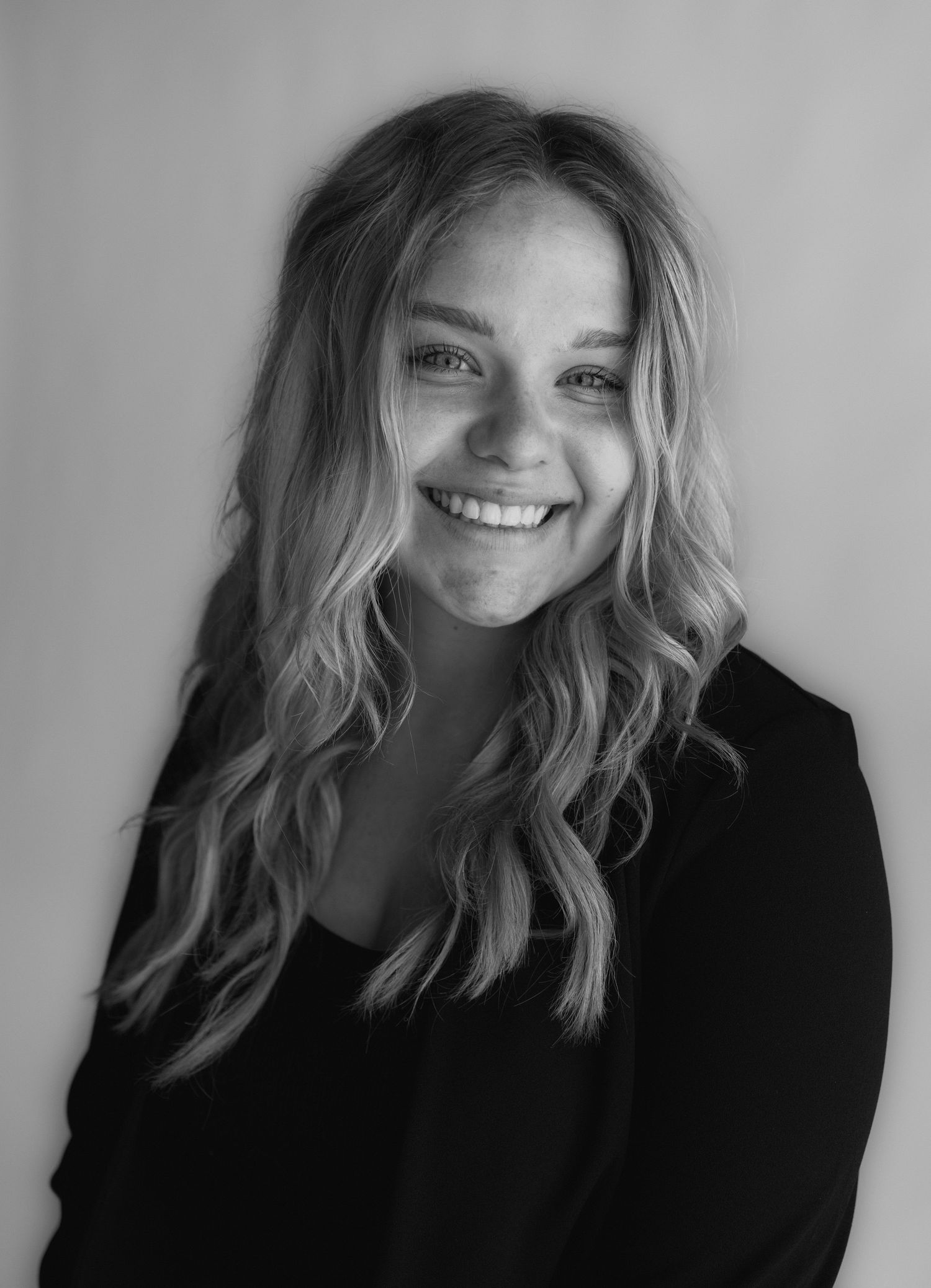 Professional black and white portrait of person with wavy blonde hair smiling while wearing a dark blazer against light background.