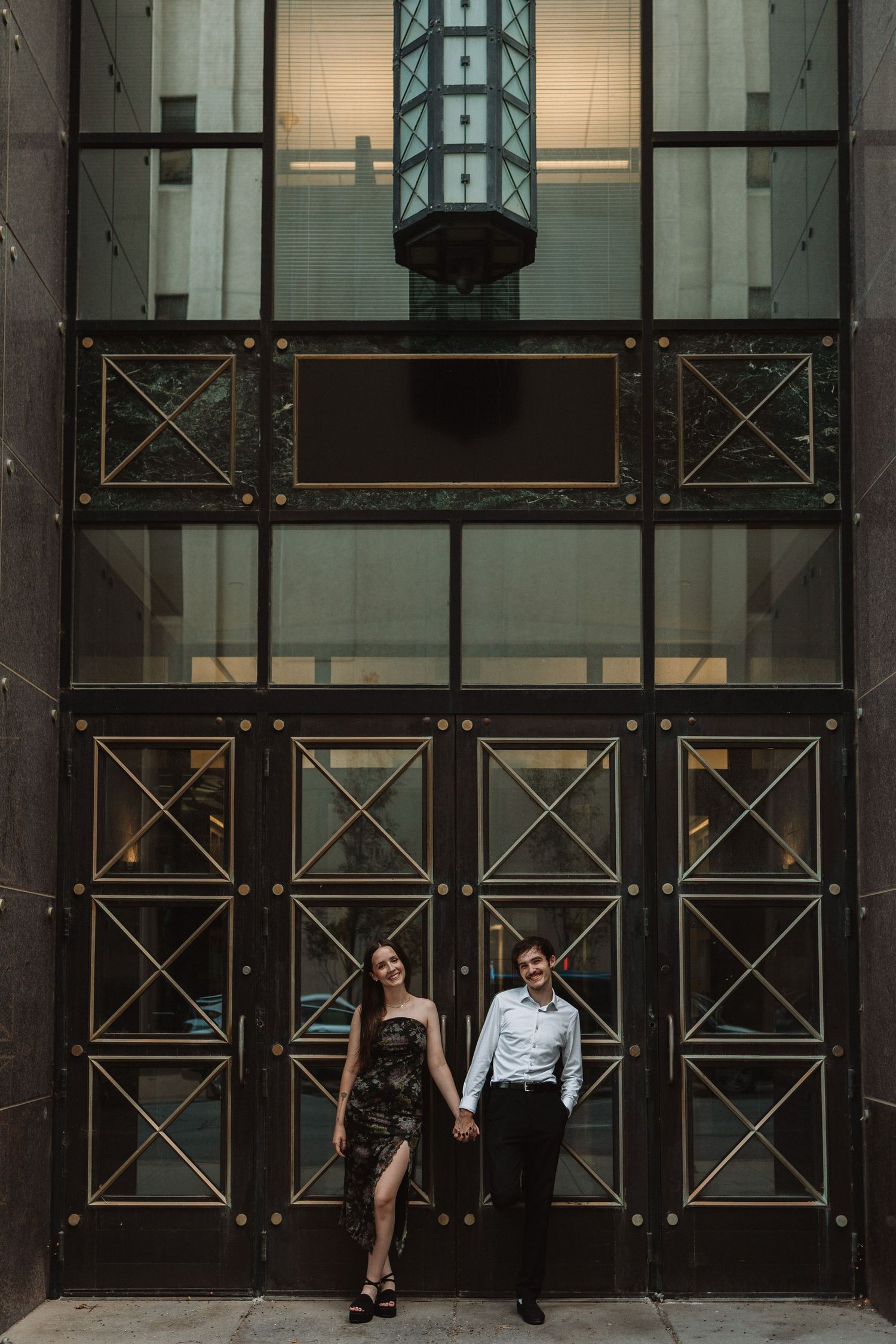 A couple stands in front of an industrial metal door with geometric patterns and art deco architectural details.