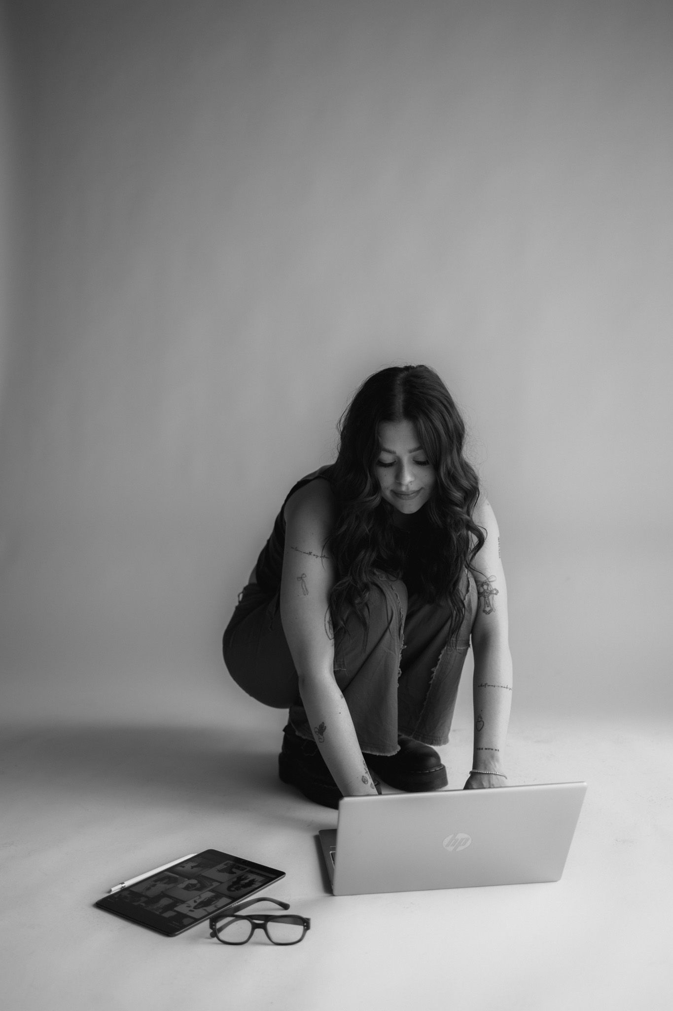 A person in dark clothing sits cross-legged on white floor working on laptop with glasses nearby in black and white photo.