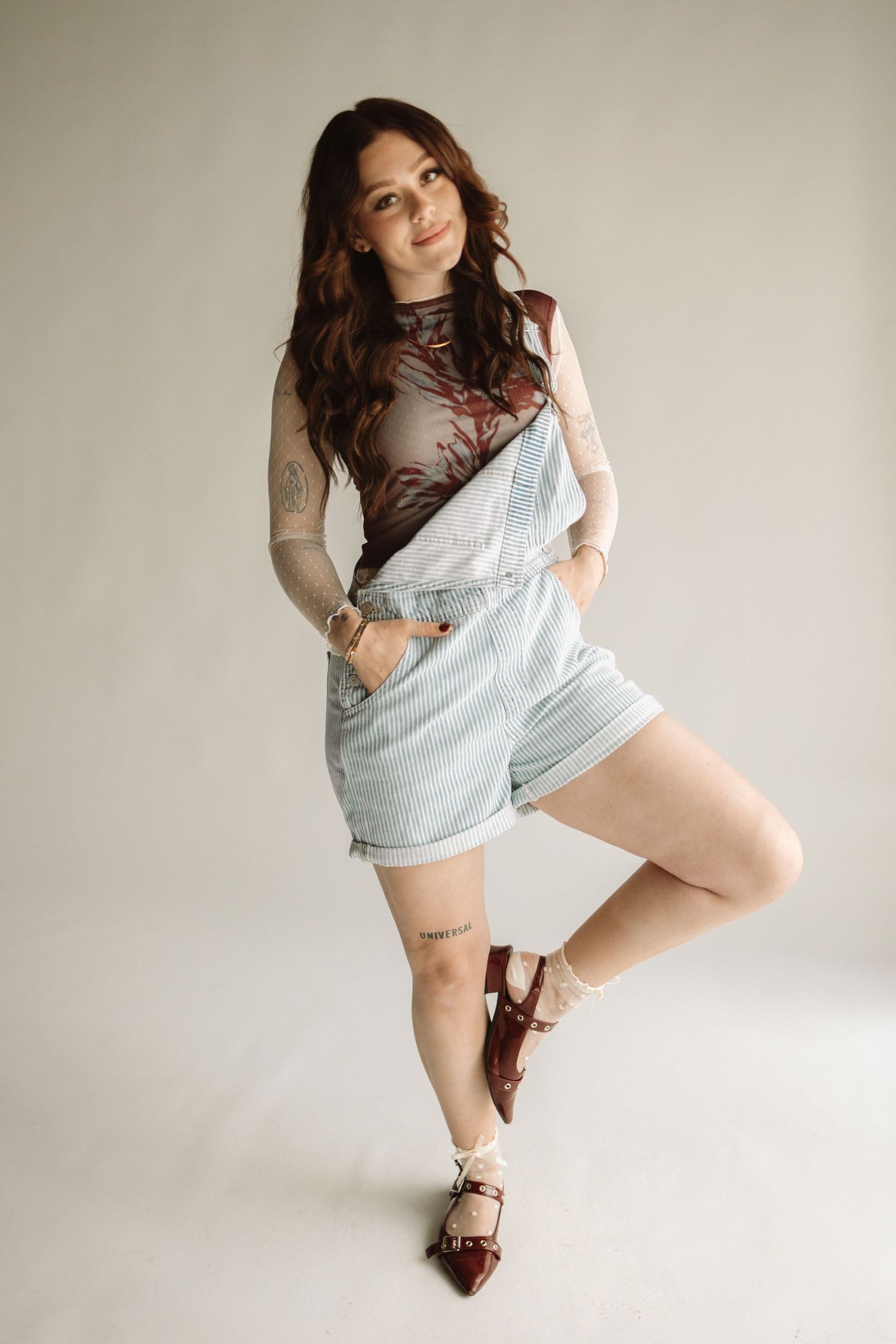 A person poses in light denim overalls and a brown mesh top against a neutral background with sandals.
