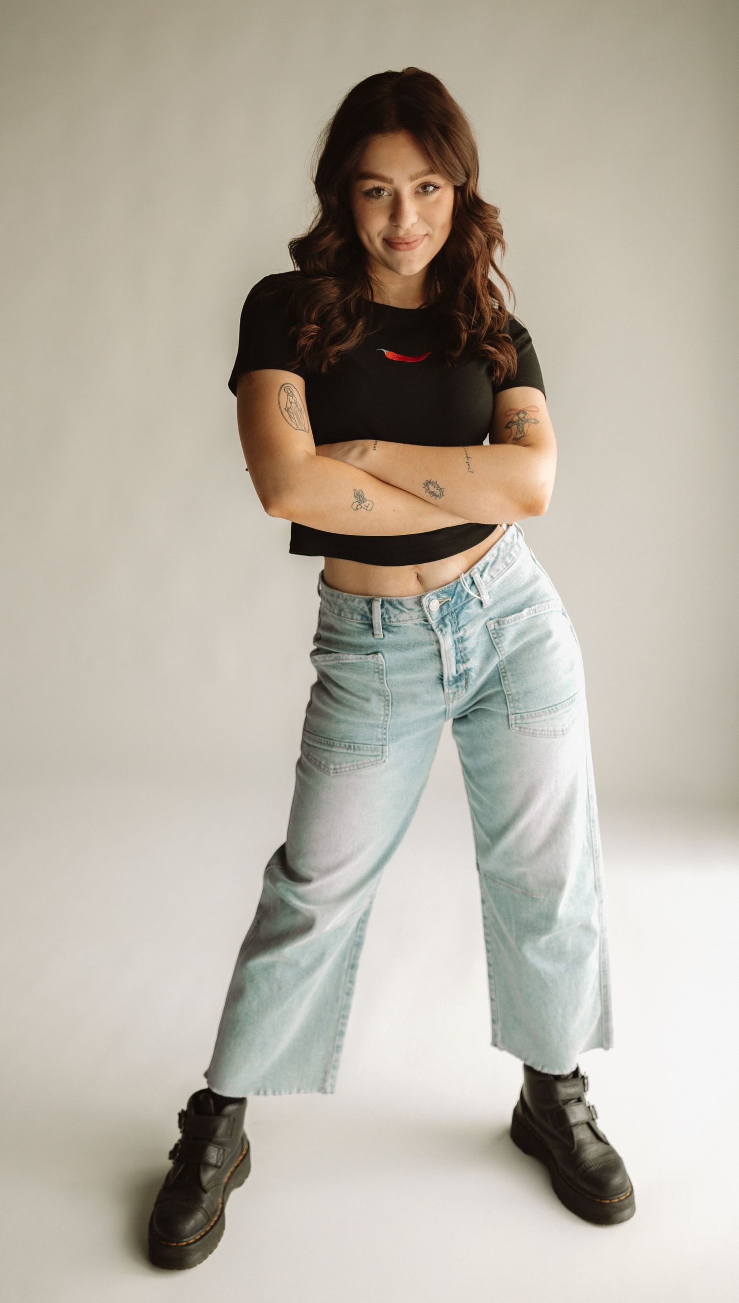 Person in black crop top and light wash baggy jeans poses with crossed arms against neutral background.