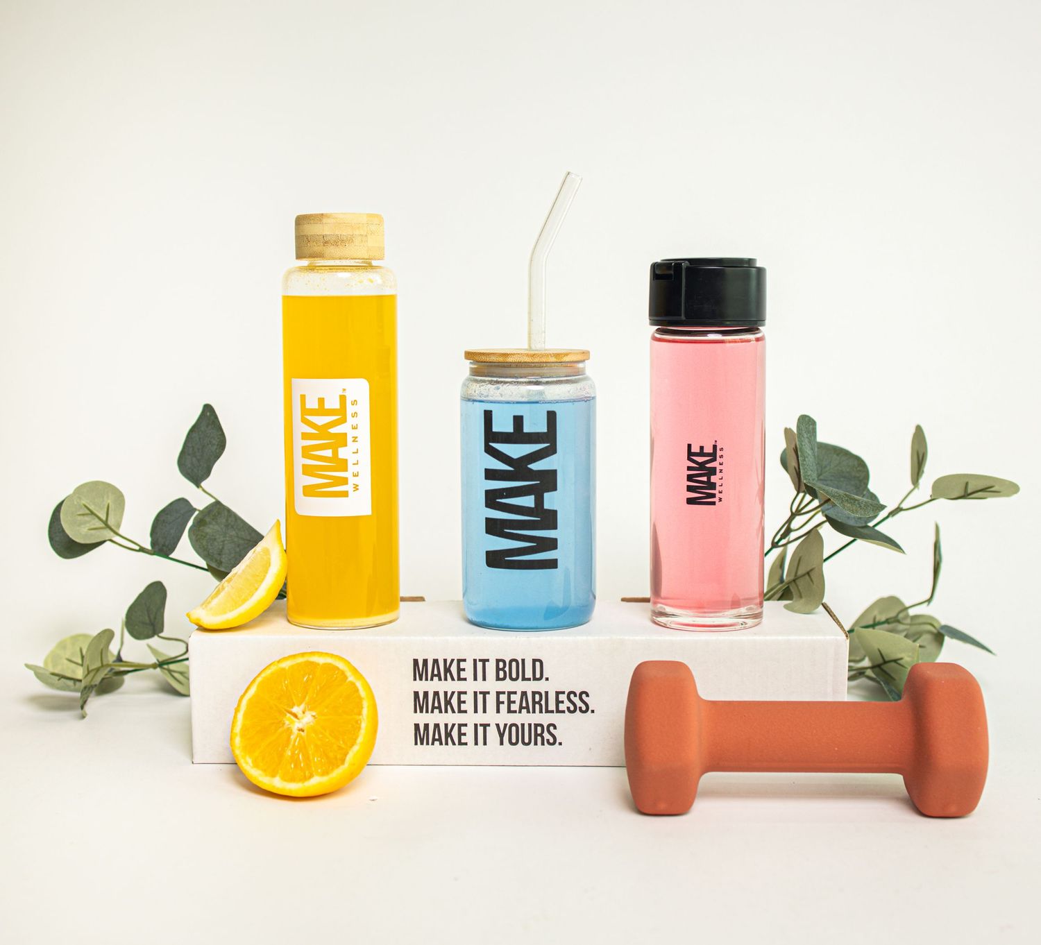 Colorful glass bottles with citrus fruits and eucalyptus leaves arranged in a minimalist product photography setup.