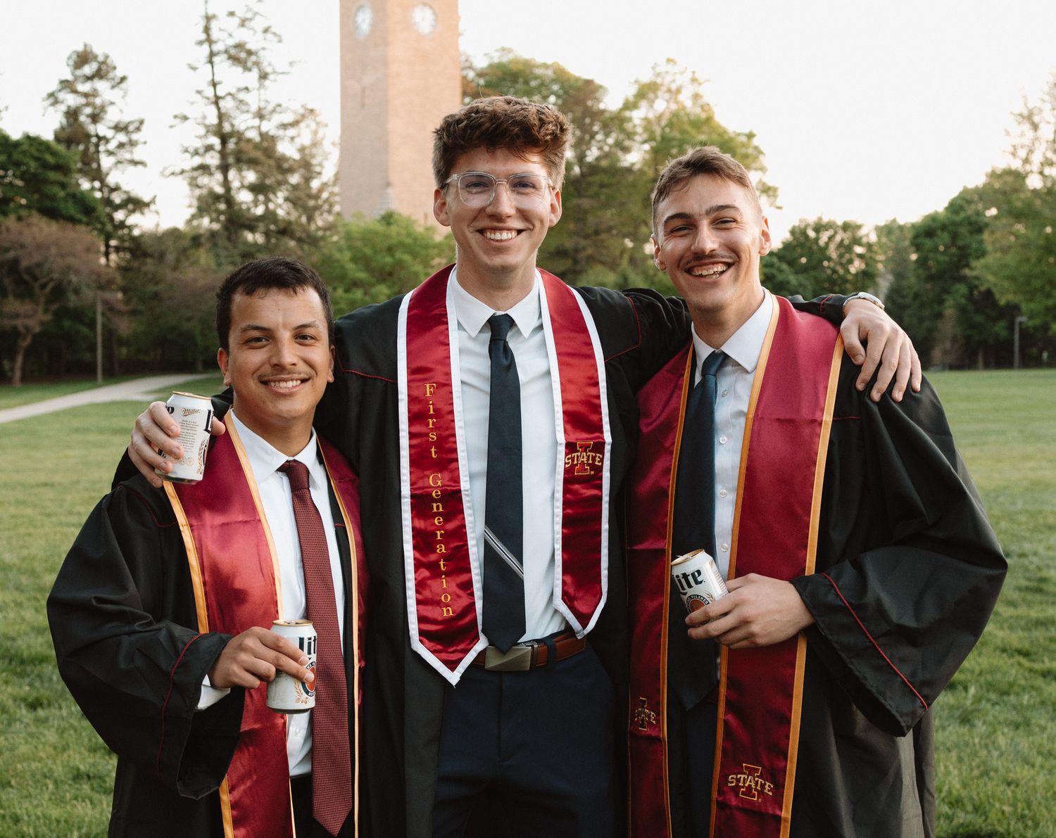 A joyful group of graduates in black robes and red stoles celebrate together outdoors with a clock tower visible in the background.