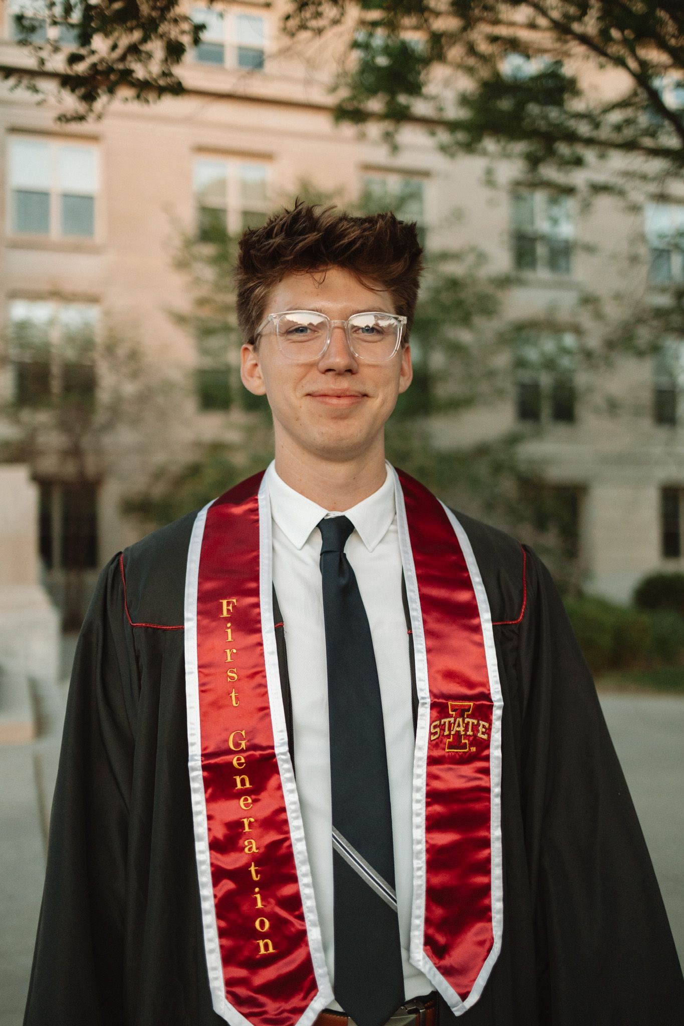 Graduate wearing red and black commencement attire standing outside academic building during graduation ceremony.