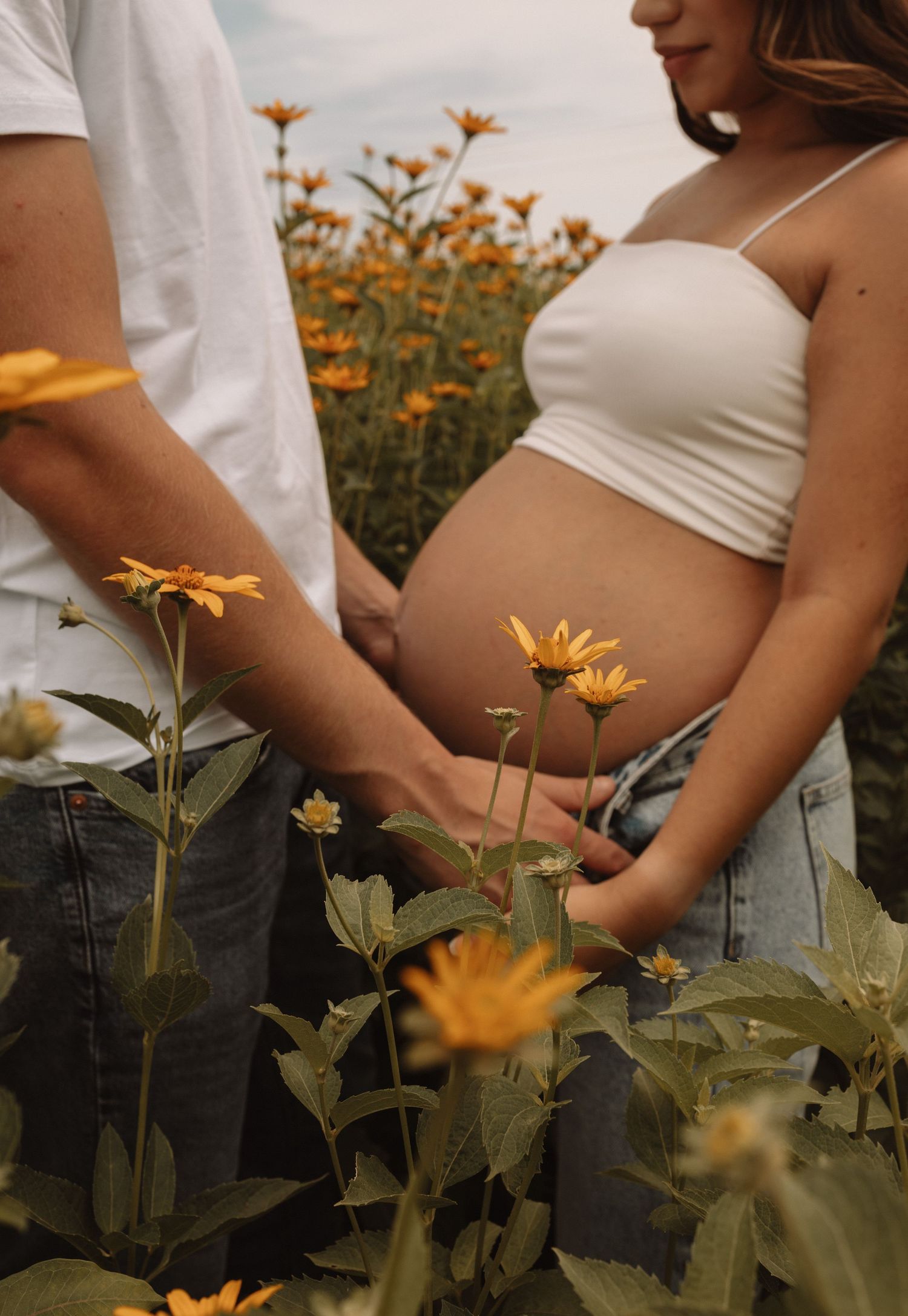 A pregnant woman in white crop top stands in a field of blooming yellow wildflowers on a cloudy day.