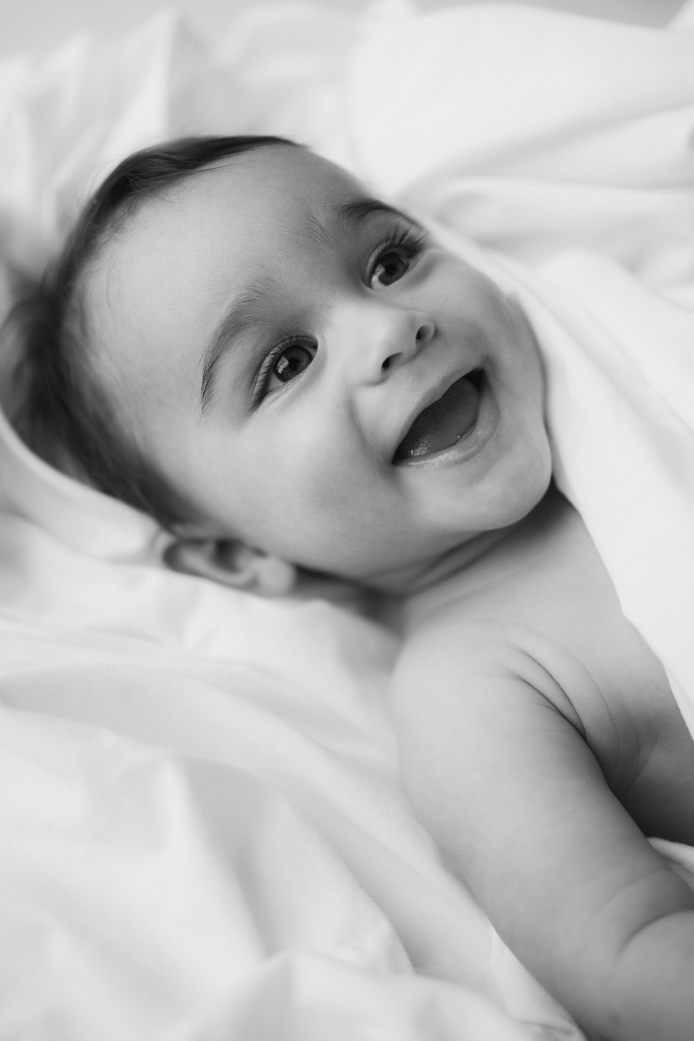 Close-up portrait of a joyful baby with bright eyes lying on white fabric.